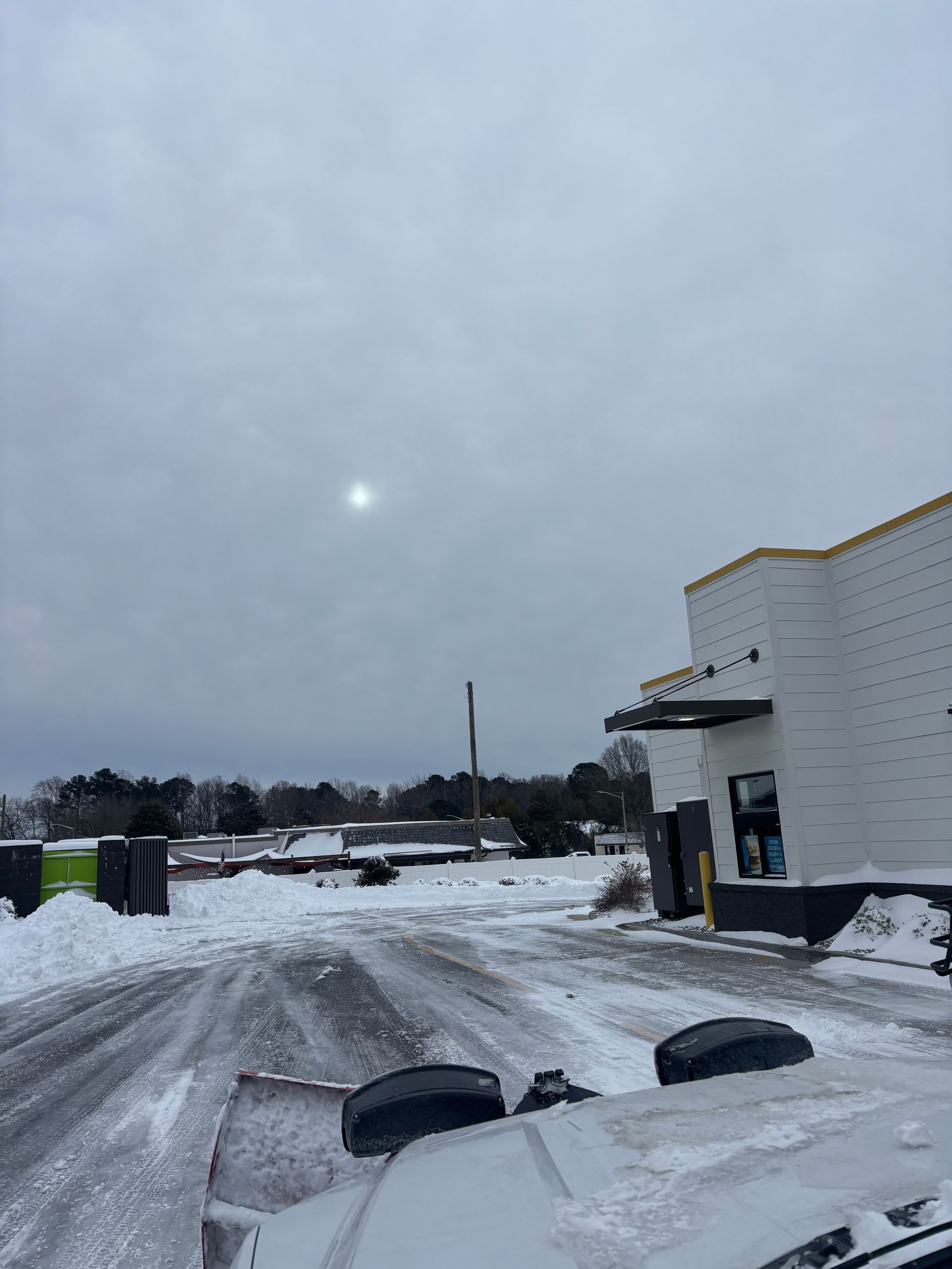 Snowy parking lot beside a building under a gray sky, with tire tracks and the sun visible near the horizon