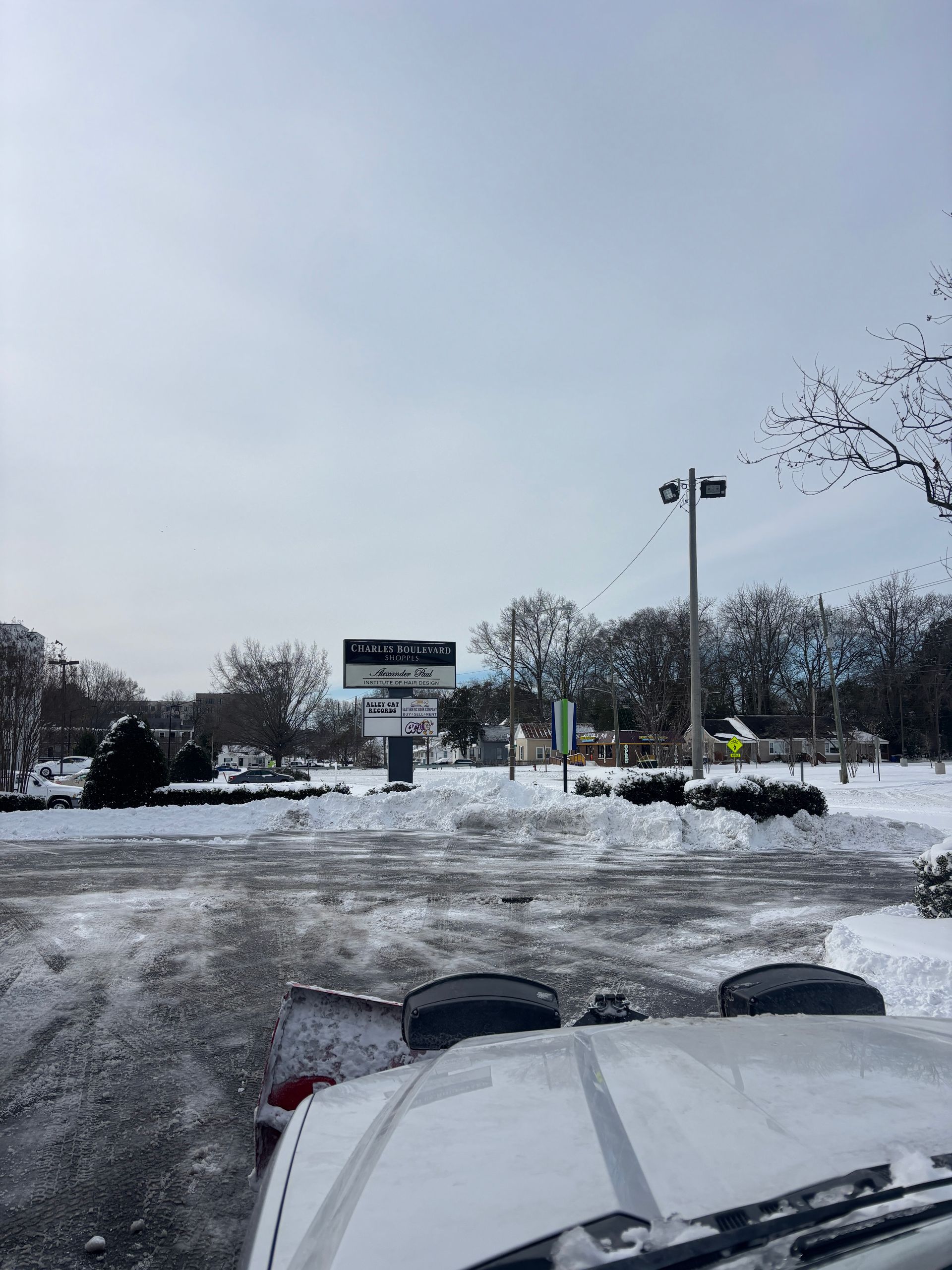 Snowy parking lot with a sign and lampposts, seen from inside a vehicle with snow-covered hood.