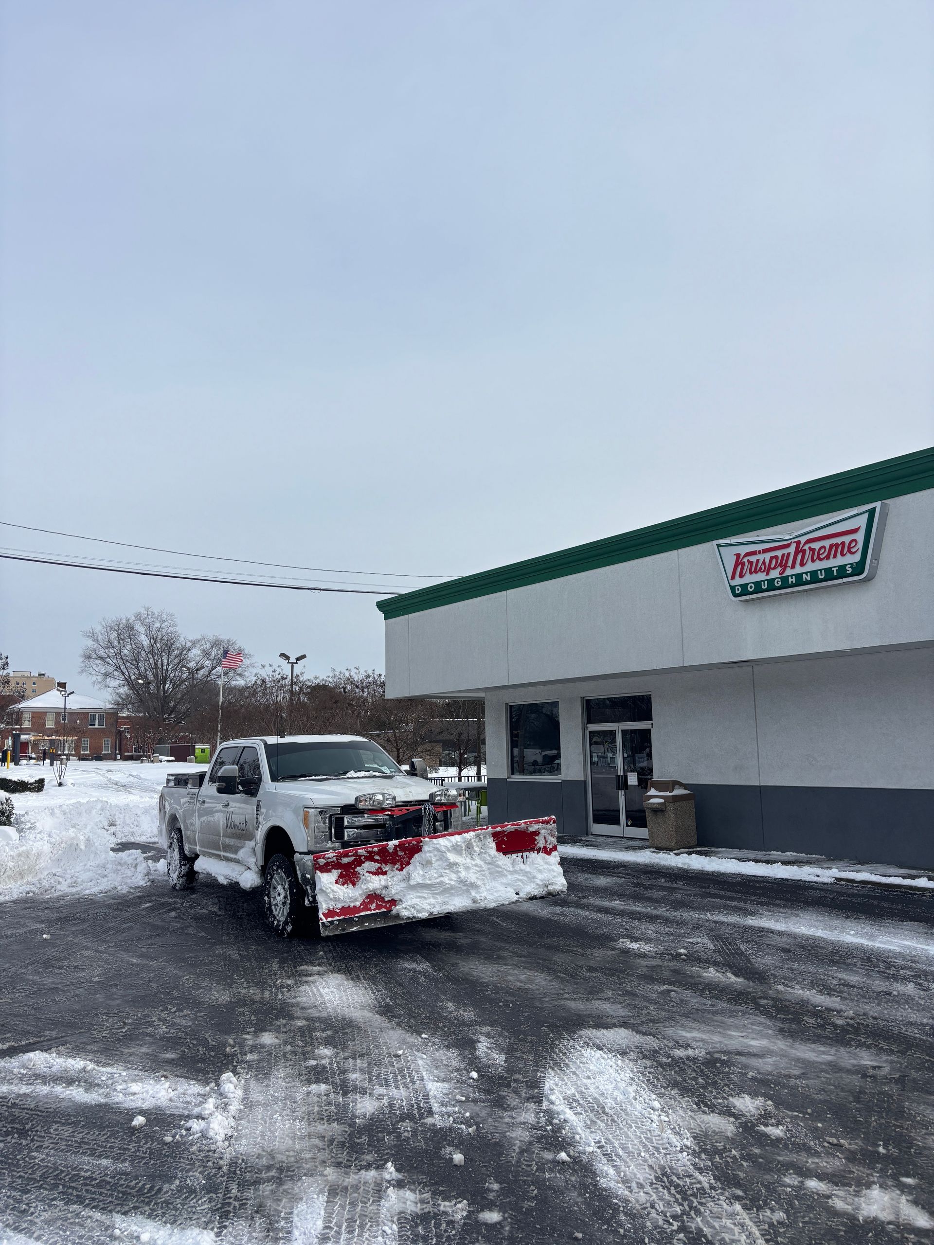 Snowplow clearing a snowy parking lot beside a store with a green-trimmed roof