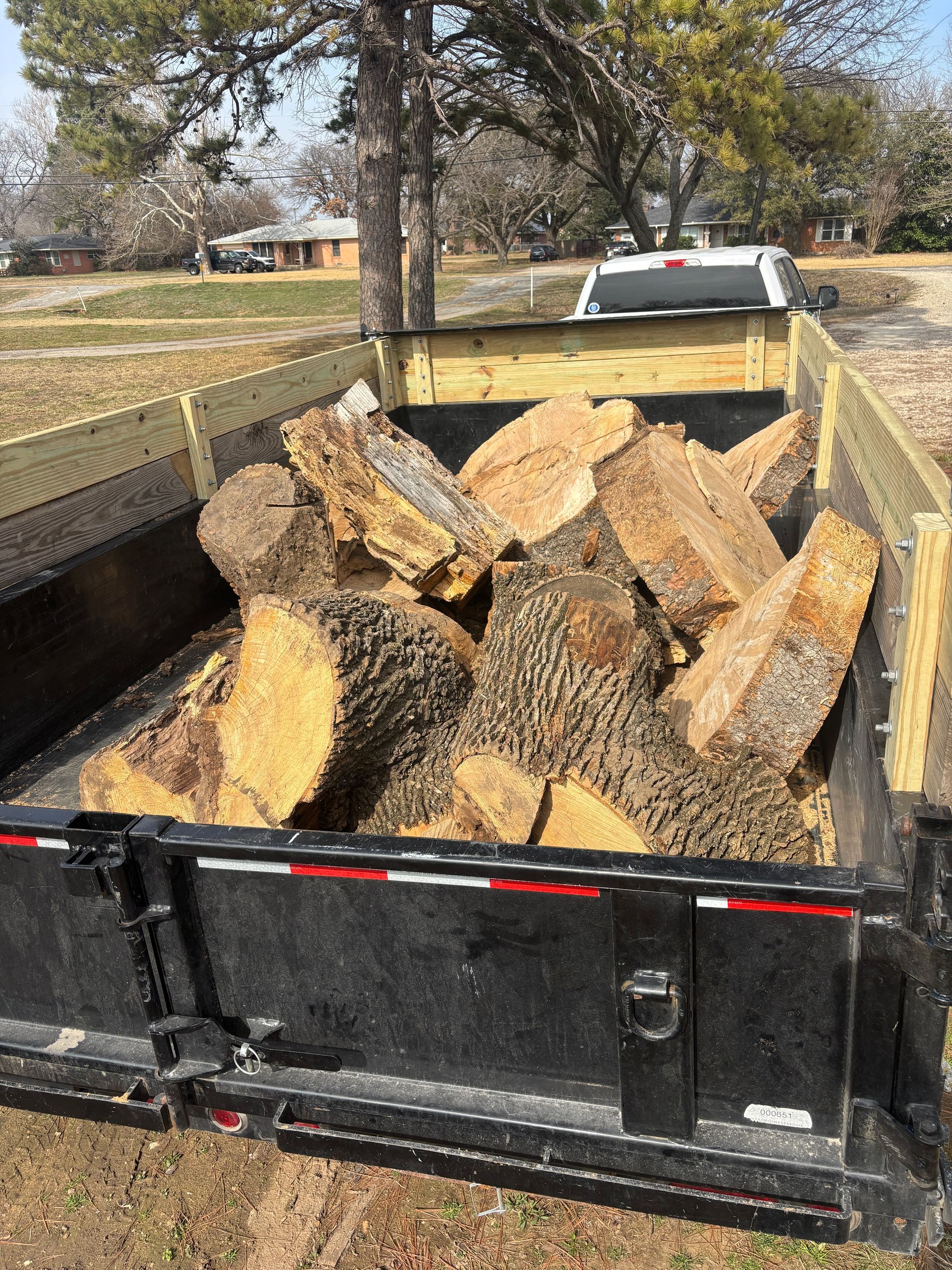 Black trailer piled high with split firewood beside a white pickup in a rural yard