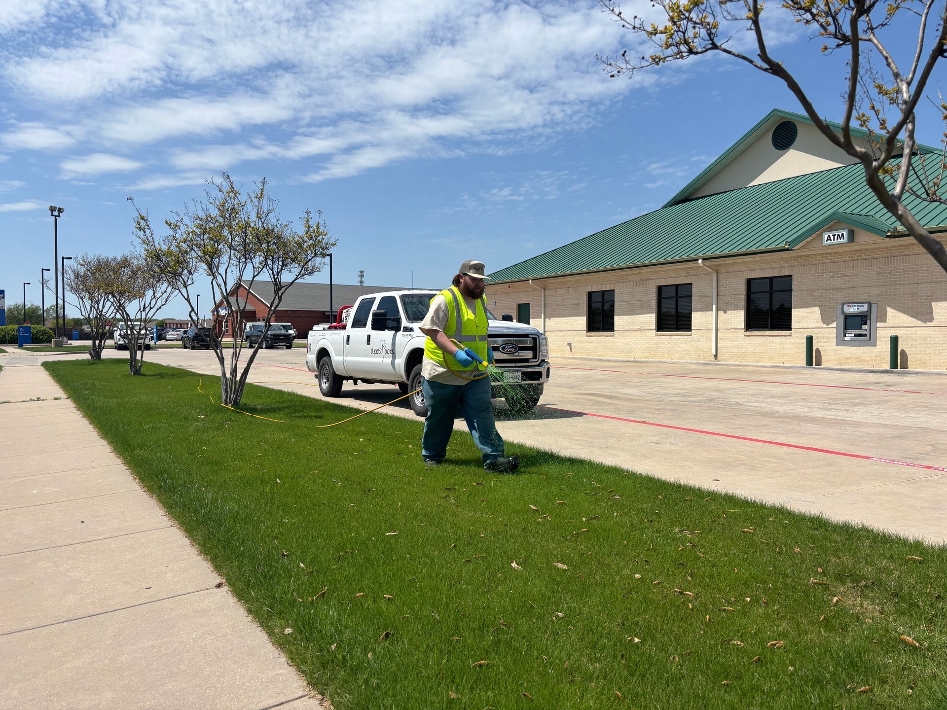 Worker in a neon vest mowing grass beside a sidewalk and building under a blue sky.