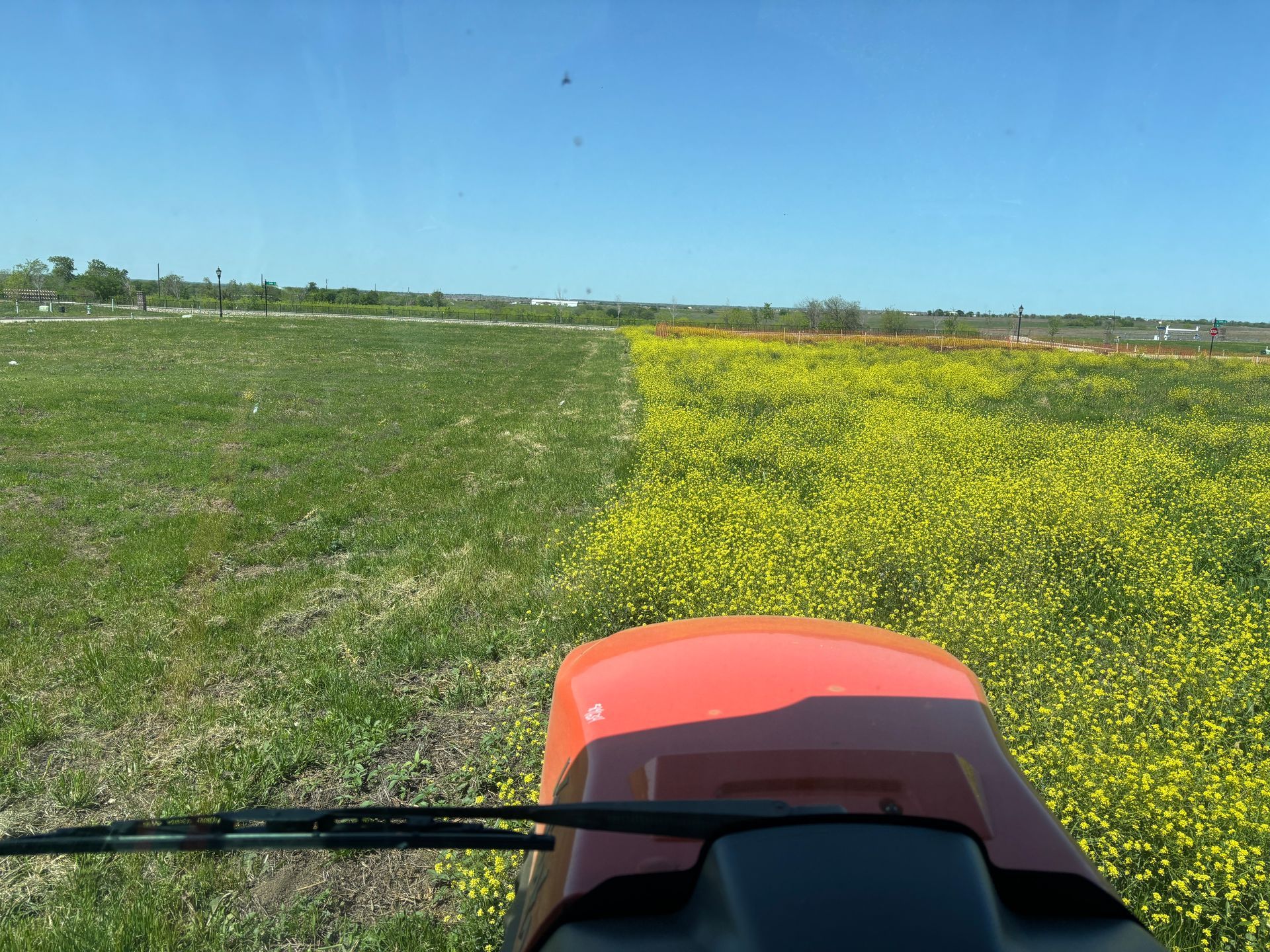 View from a red tractor crossing a green field beside a strip of yellow wildflowers under a blue sky