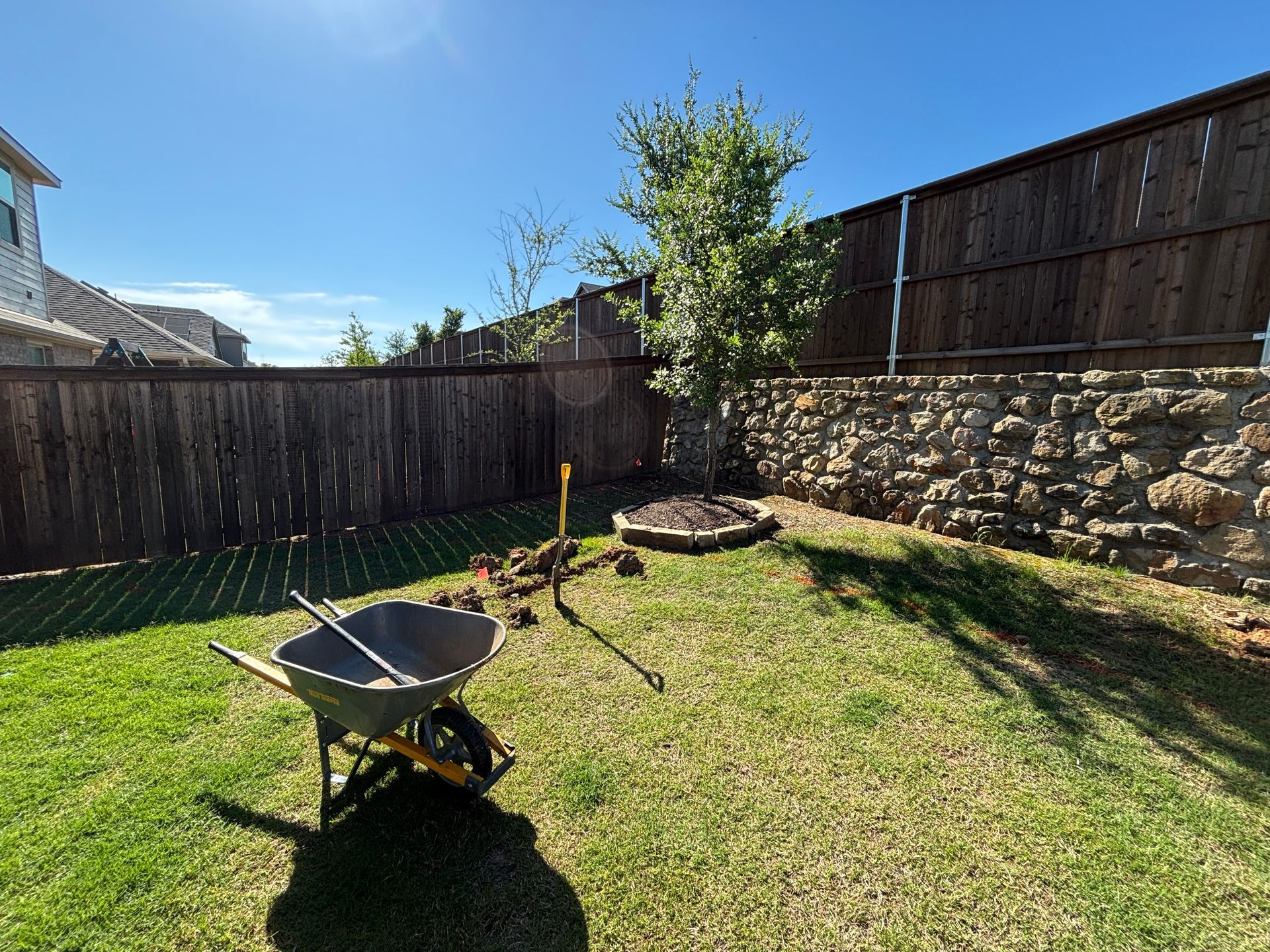 Sunny backyard with a wheelbarrow, fire pit, and wooden fence on a grassy lawn.