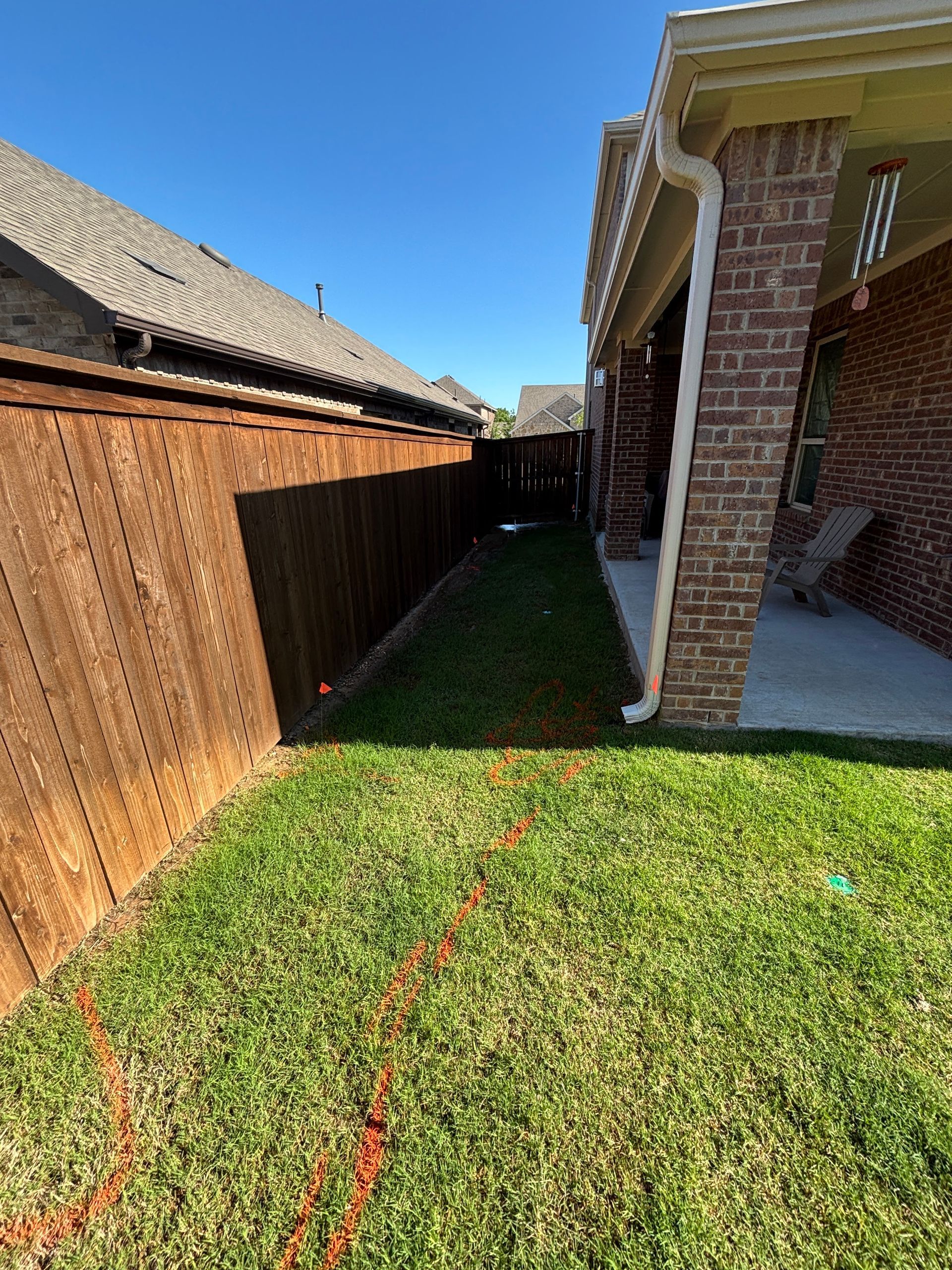 Narrow grassy side yard between a wooden fence and brick house under a bright blue sky.