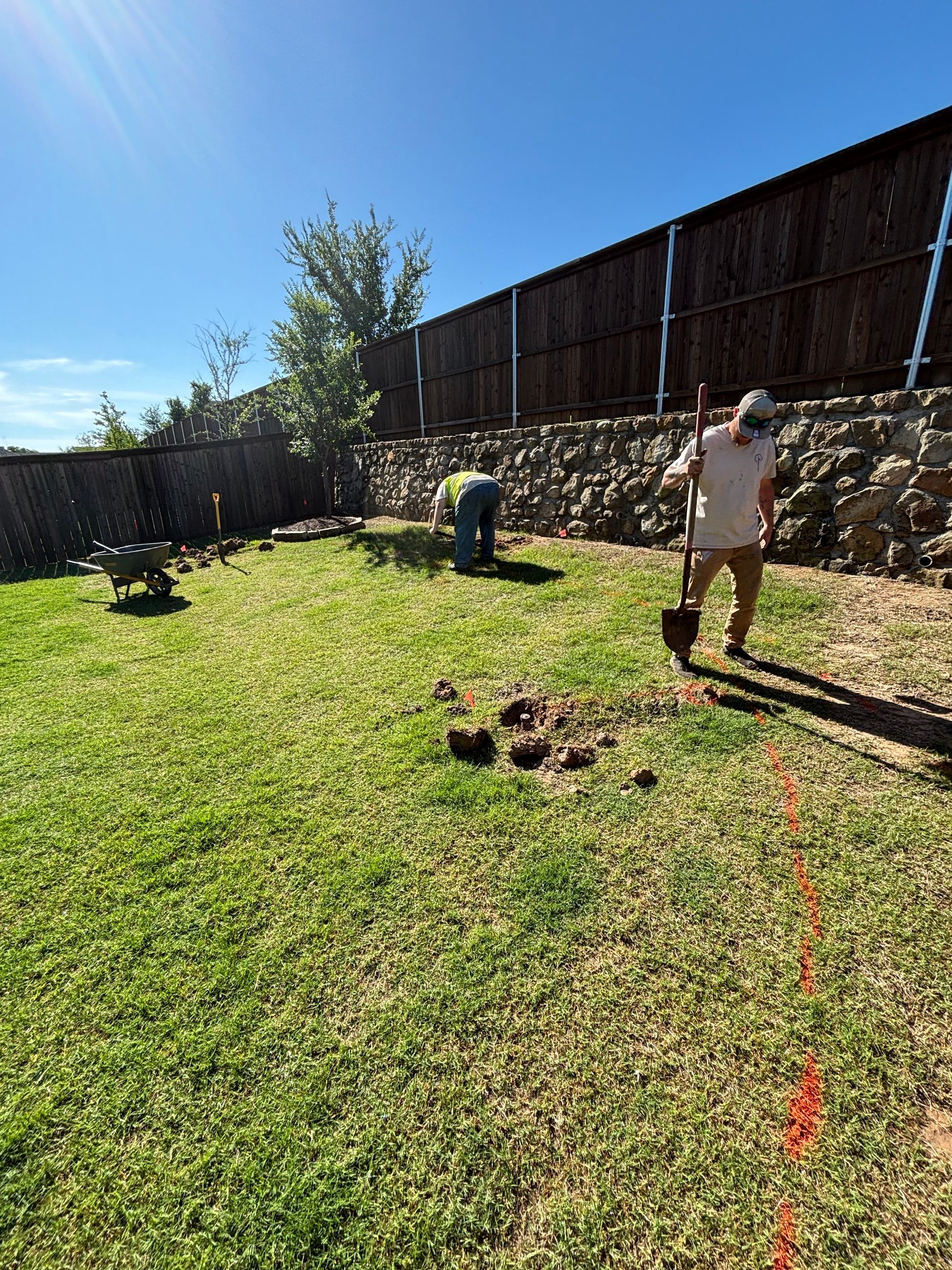 Two people working in a sunny backyard with grass, a fence, and a long orange hose.