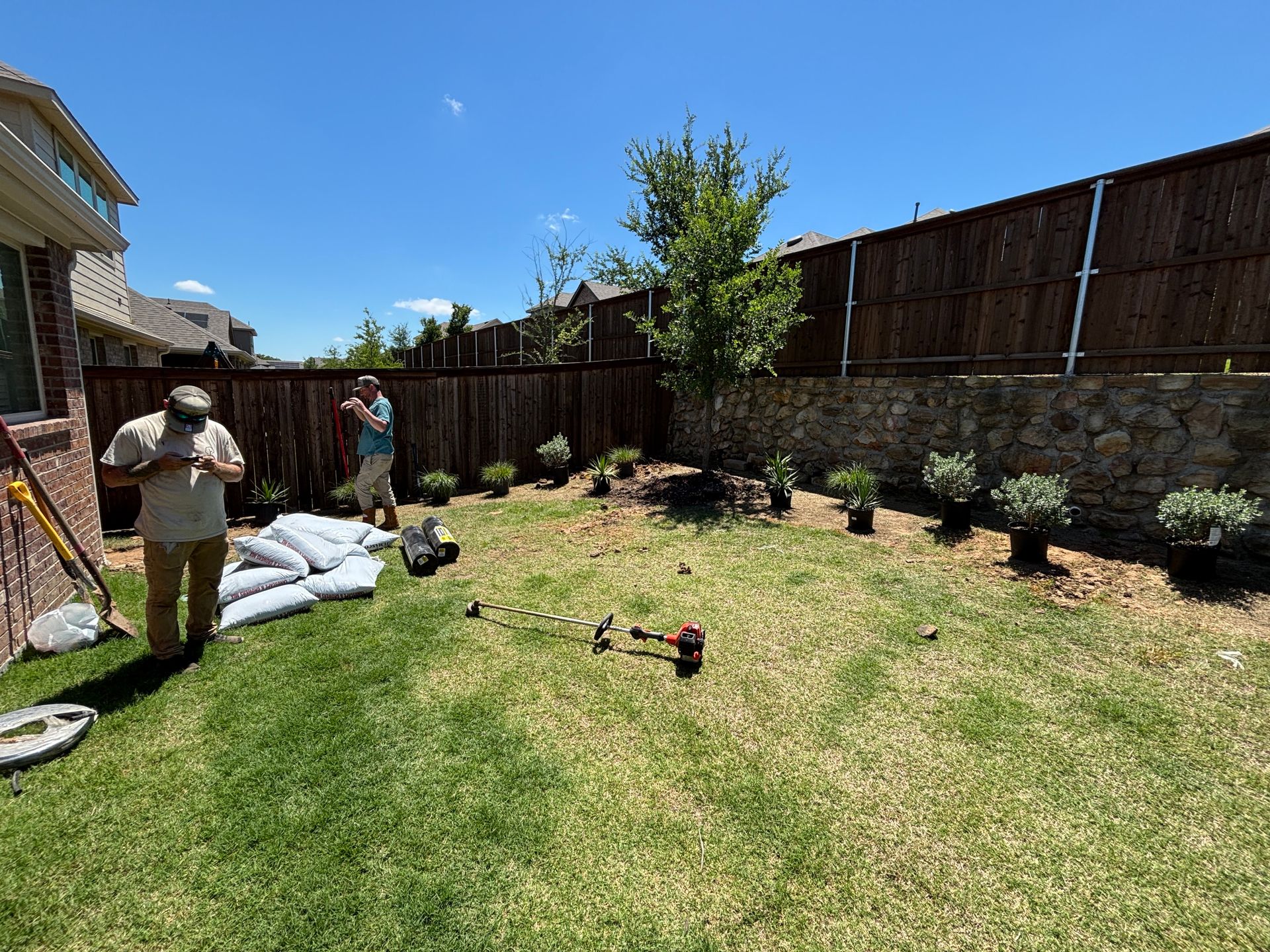 People working in a backyard garden beside a wooden fence and stone wall on a sunny day