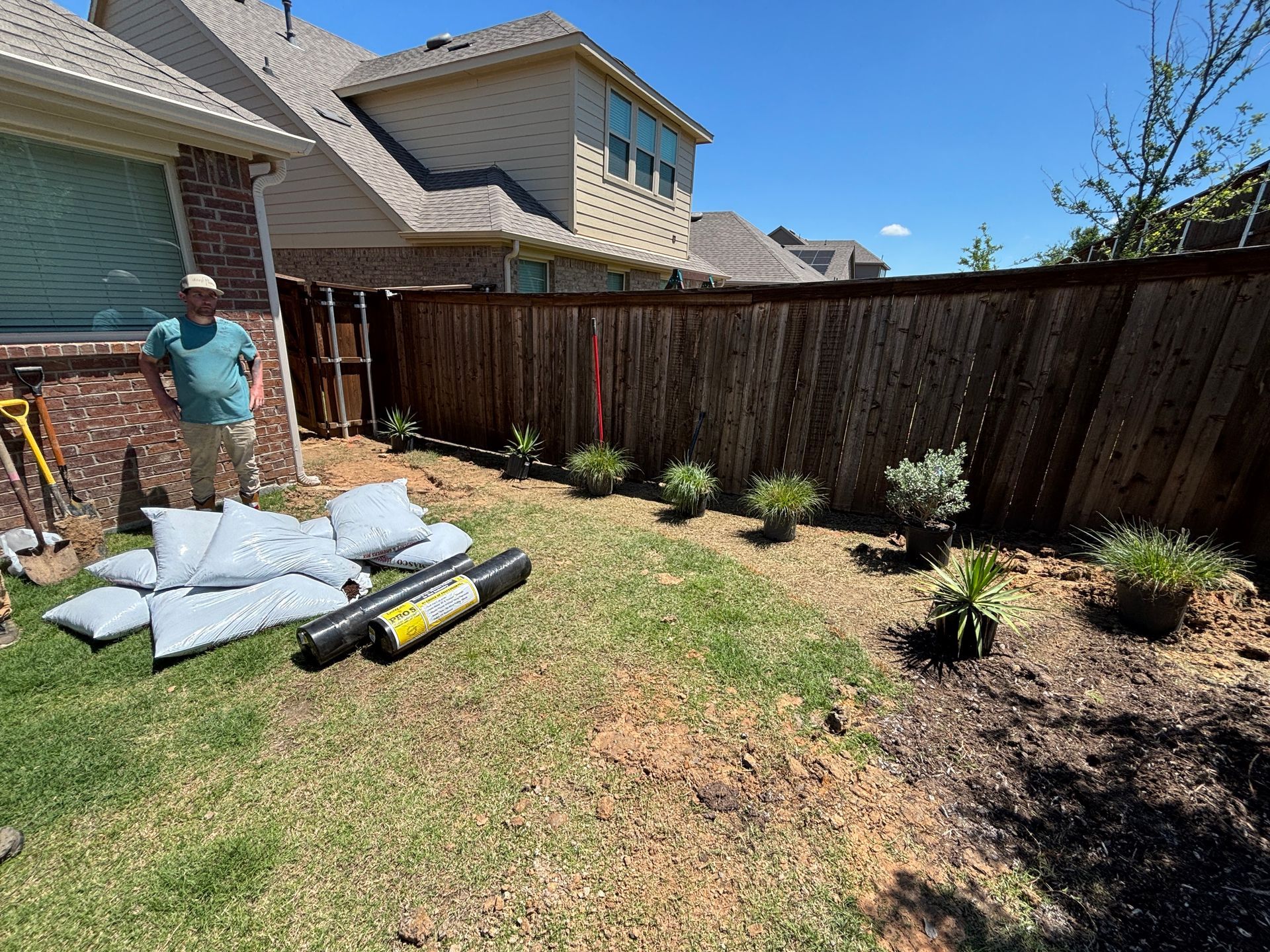 Worker in teal shirt by backyard fence, with landscaping materials and newly planted shrubs.