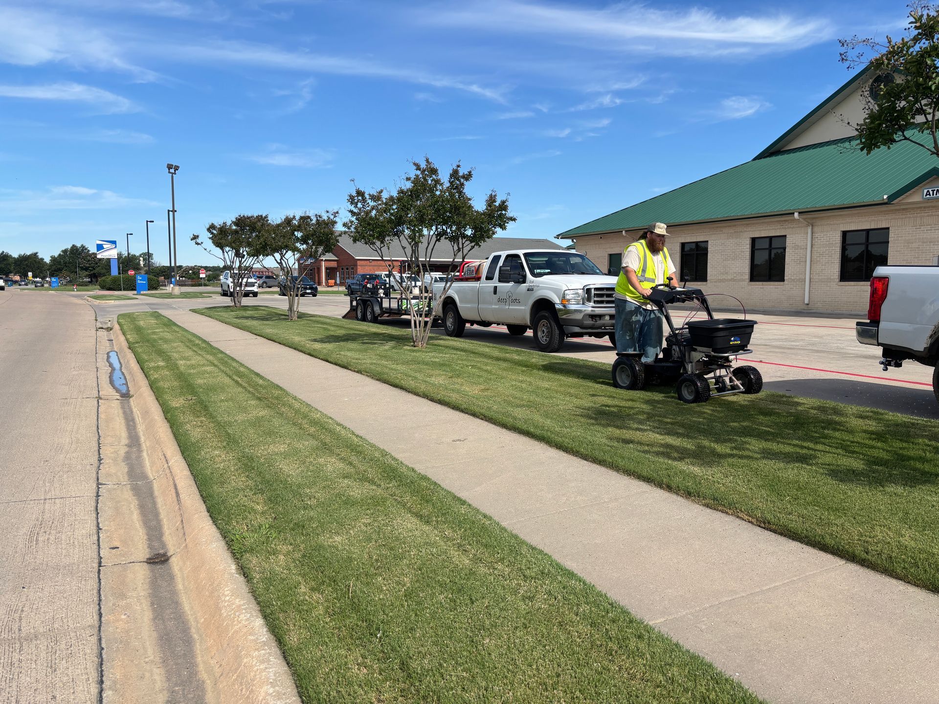 Person on a standing lawn mower next to vehicles on a sidewalk near a building with a green roof.