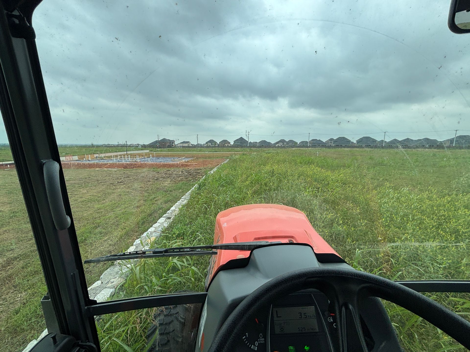 View from a tractor cab over green field and cloudy sky, with orange hood in foreground