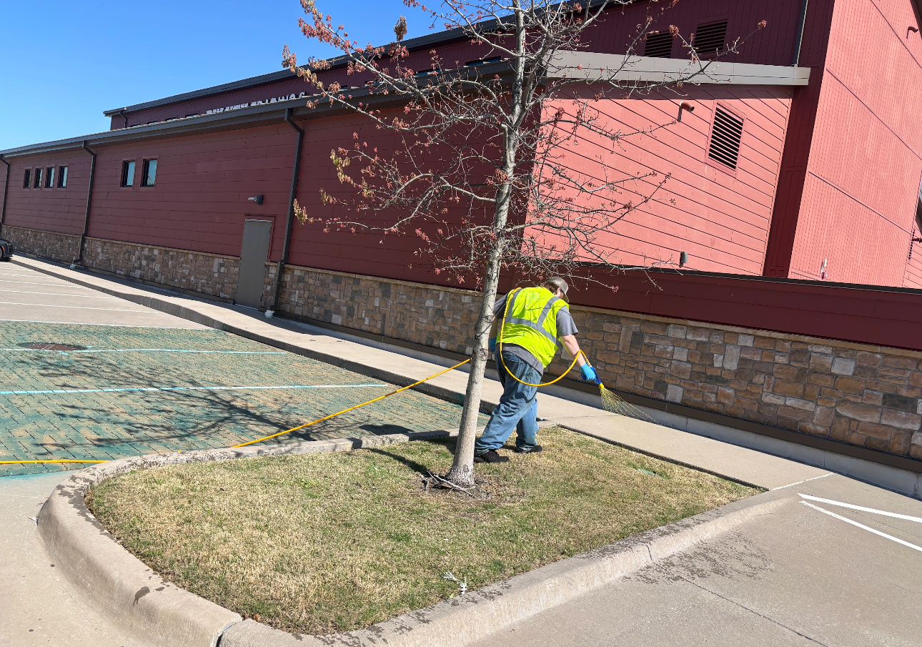 Worker in a neon vest trimming grass beside a red brick building on a sunny day