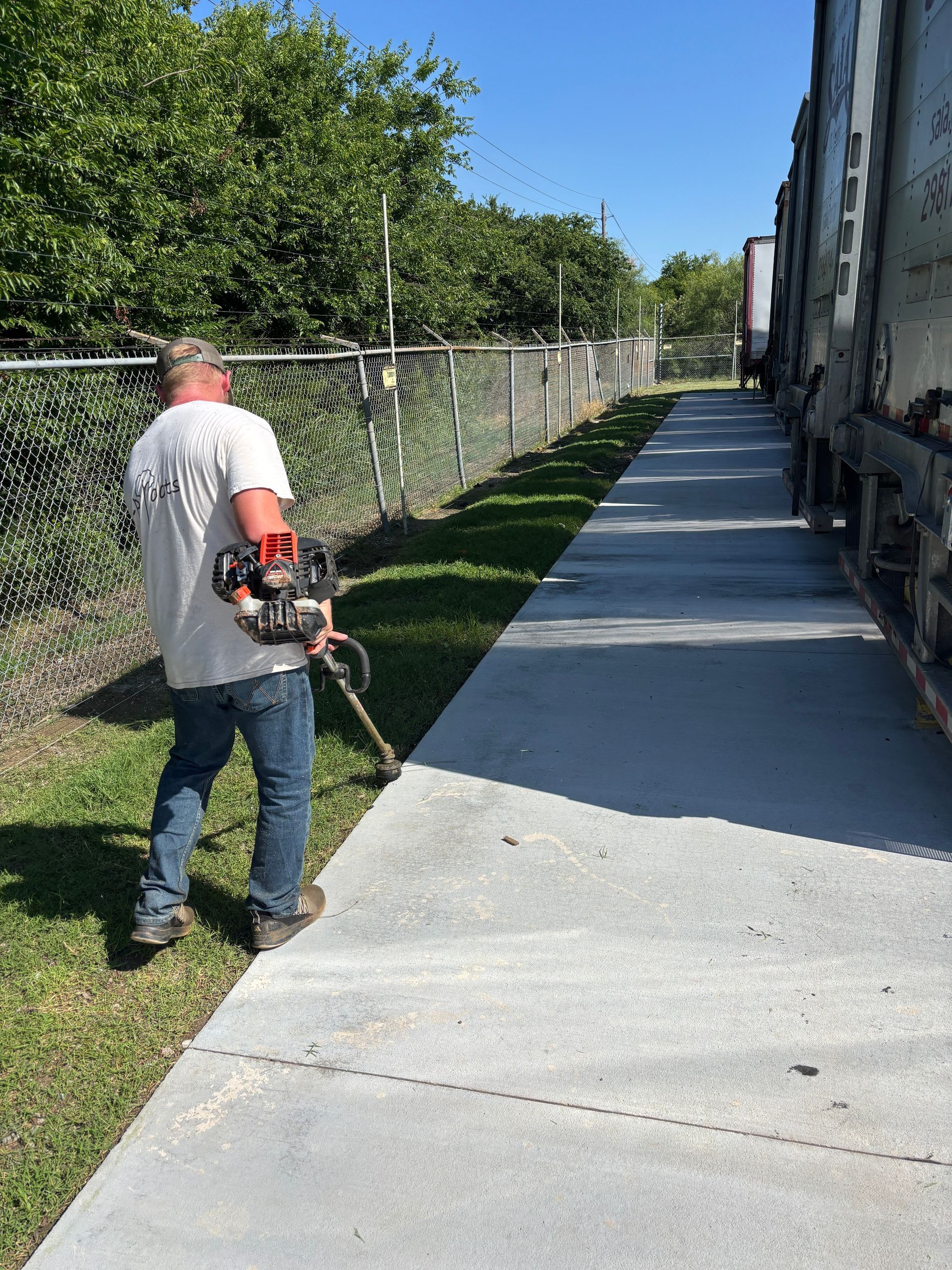 Worker using a string trimmer along a sidewalk beside a building and chain-link fence