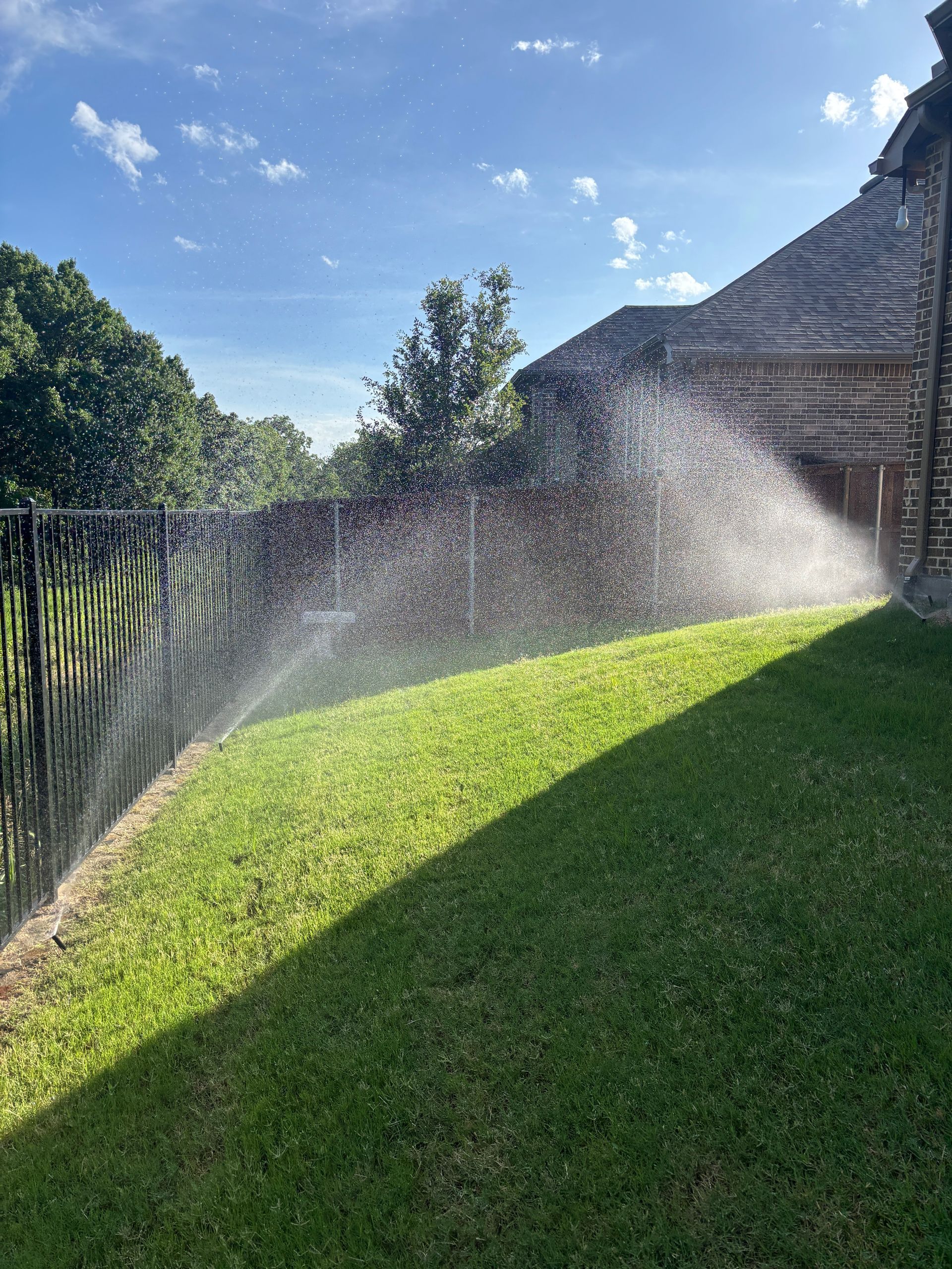 Sprinkler mist over a sunlit green lawn beside a wooden fence and house wall