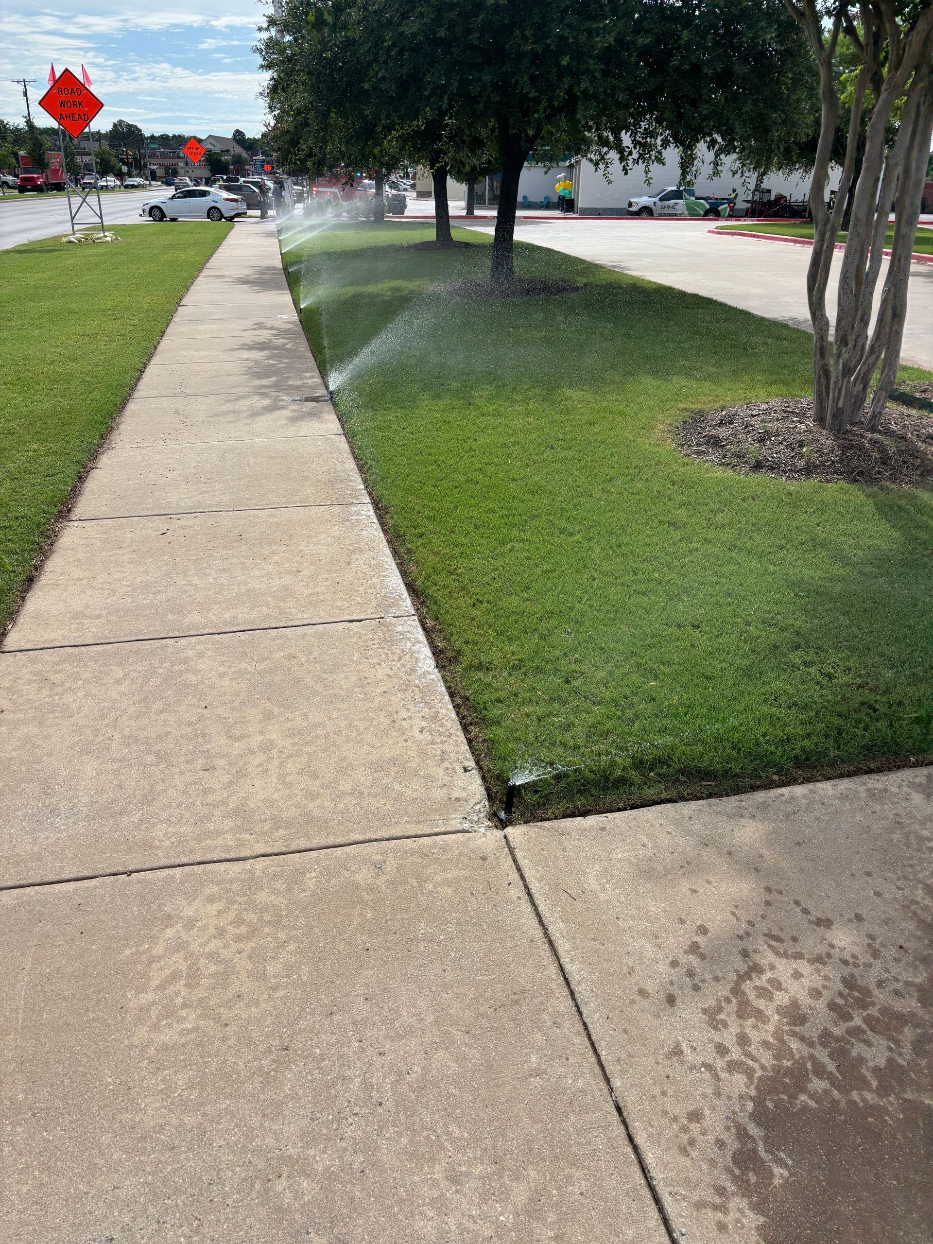 Sidewalk beside a grassy lawn with sprinklers spraying water near trees and a road