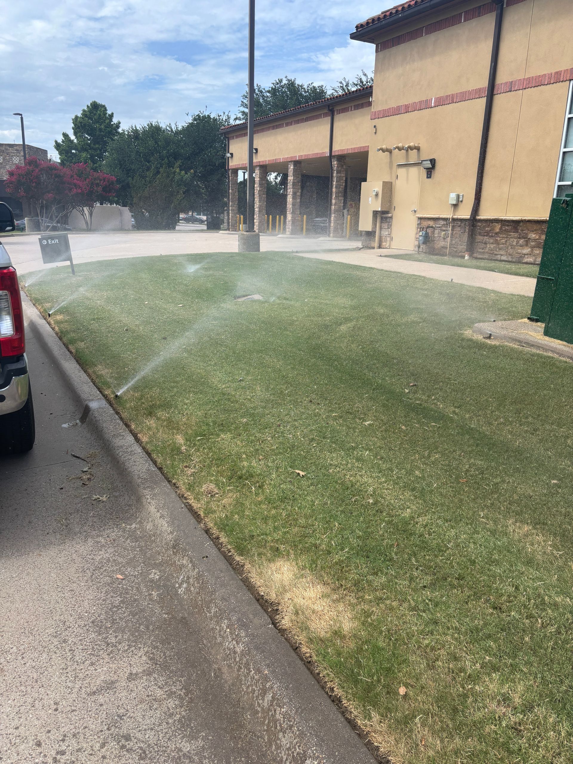 Sprinklers watering a grassy slope beside a shopping center parking lot under a blue sky