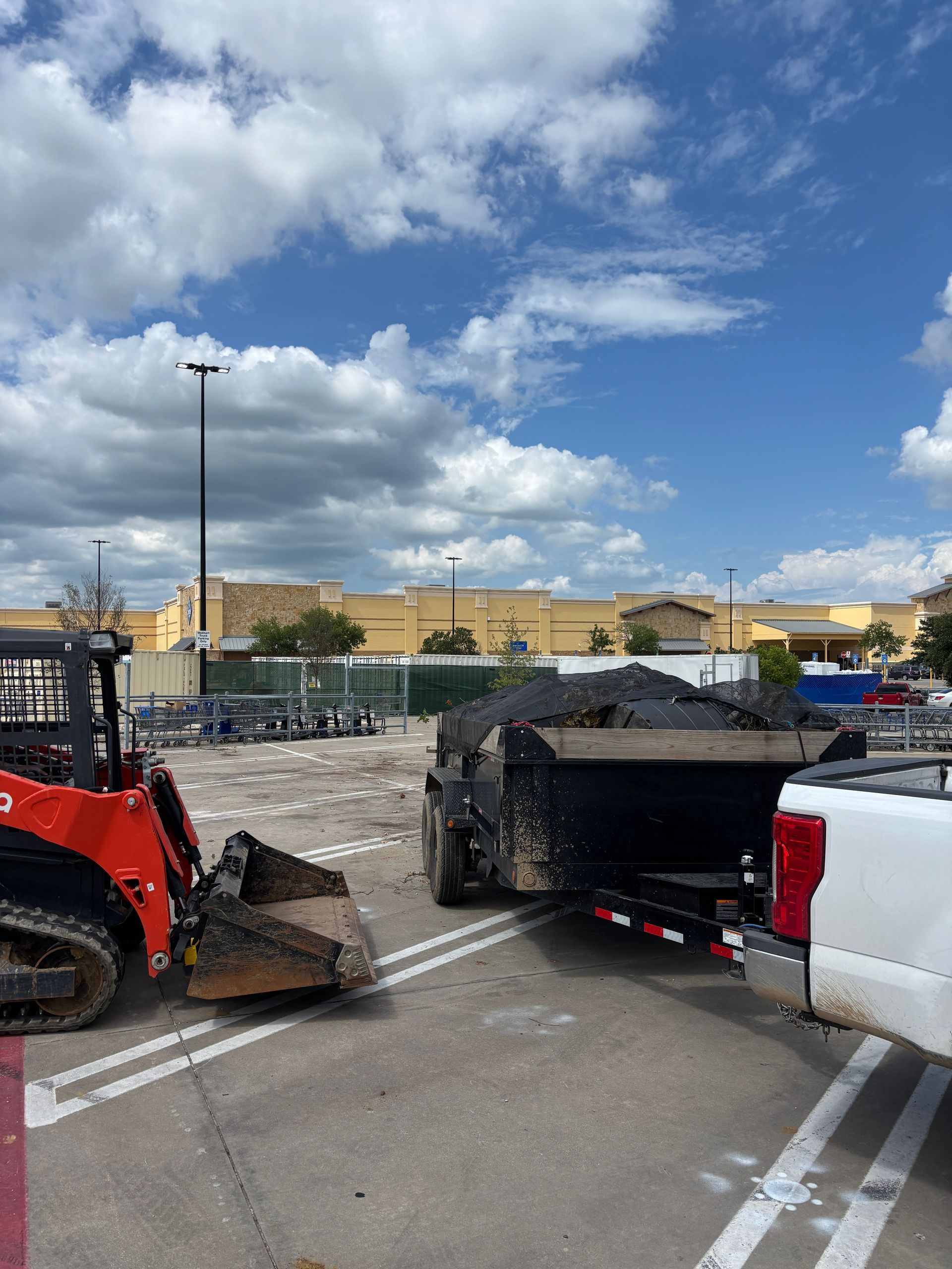 Construction vehicles in a parking lot with a black trailer and white pickup under a cloudy blue sky