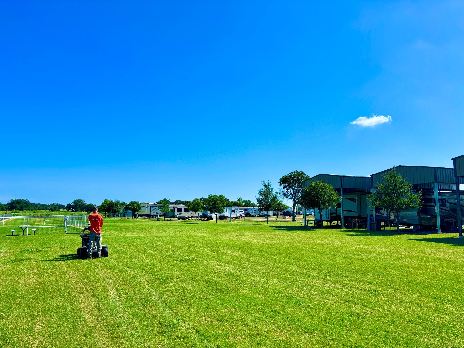 Wide green field under a bright blue sky, with a person mowing grass near the left side.