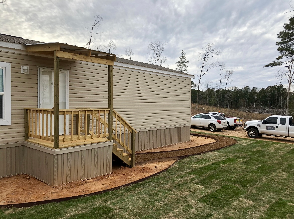 Small beige house with wooden porch and ramp on a grassy lot, with parked vehicles nearby