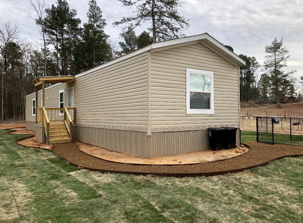 Beige tiny house with porch and ramp on a grassy lot, surrounded by trees