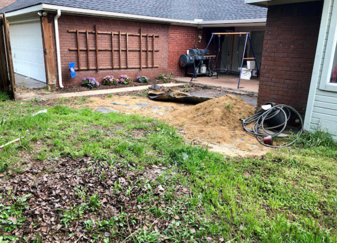Backyard with brick house, muddy yard, patio tools, and a coiled hose near the wall