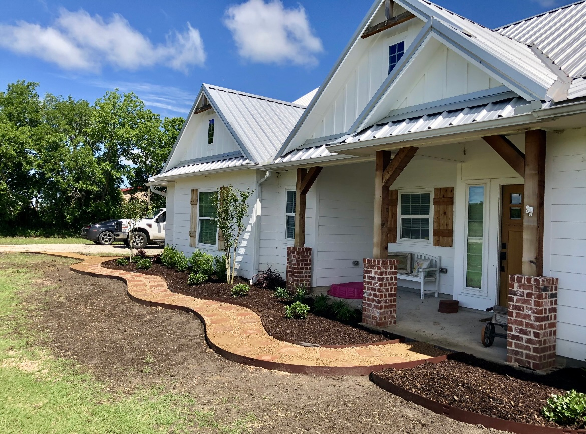 White house with front porch, brick columns, and a landscaped yard under a blue sky.
