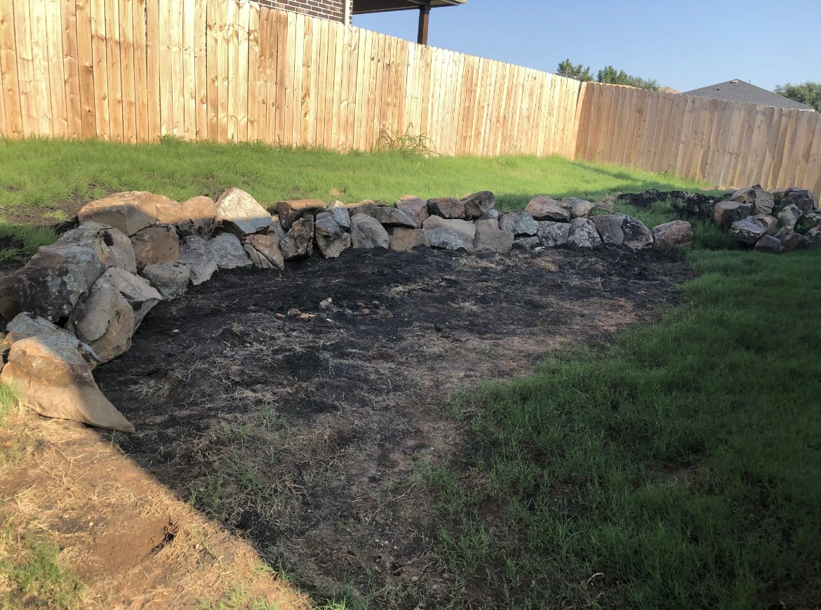 Stone-lined garden bed with bare soil beside a grassy yard and wooden fence.