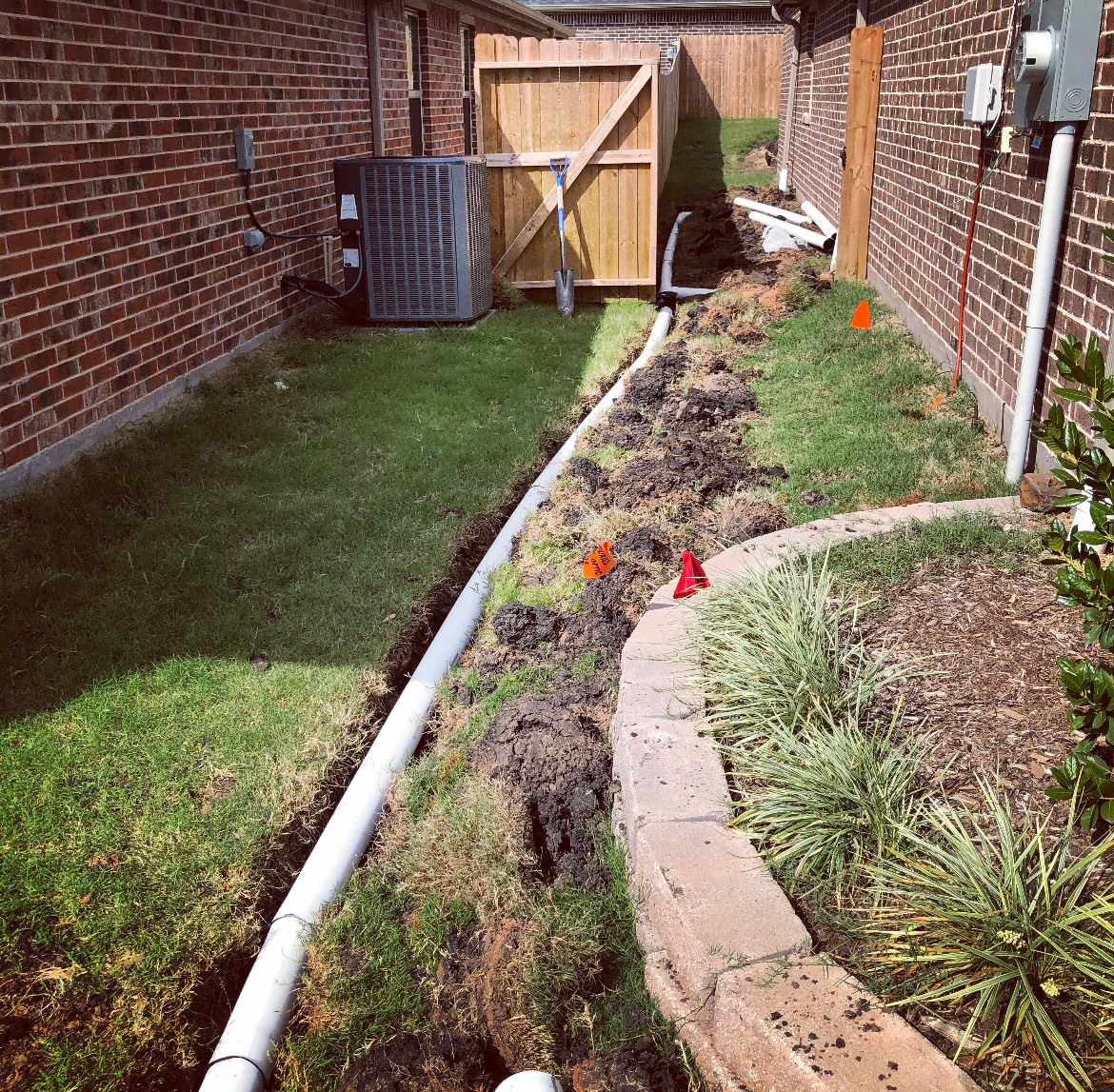 Narrow backyard trench beside brick houses with a white pipe, dirt, grass, and a wooden gate at the end