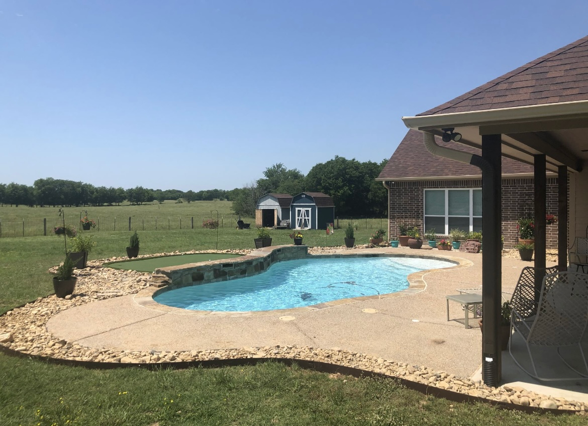 Backyard pool with stone deck beside a brick house under a clear blue sky