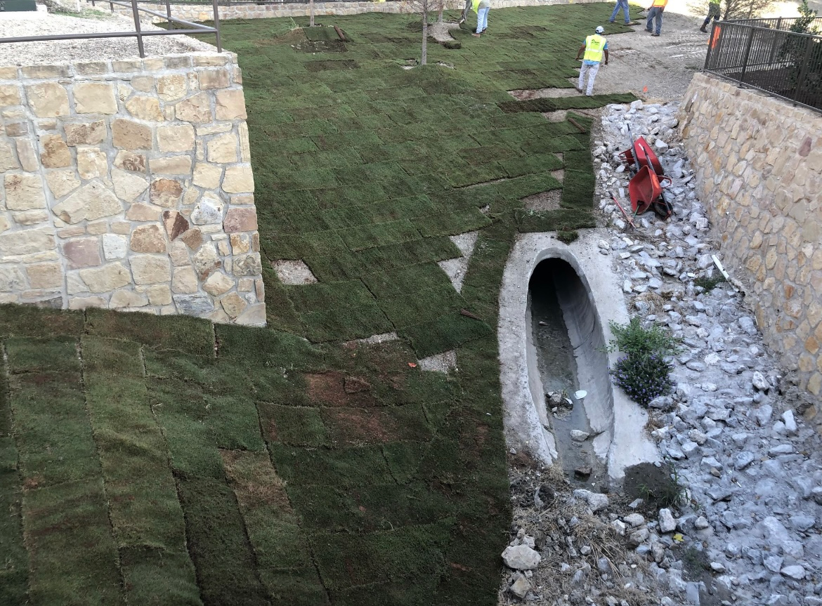 Stone drainage channel in a grassy ditch beside retaining walls, with rocks and a red object near the waterway