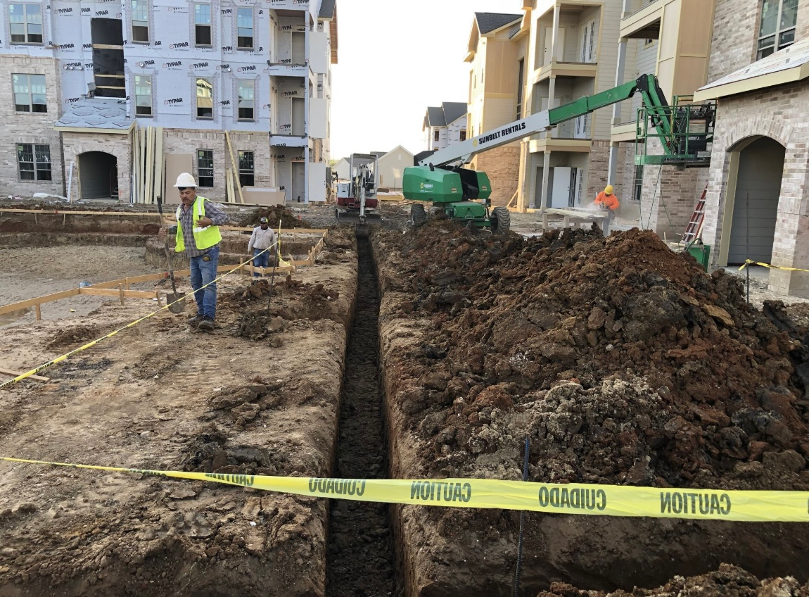 Construction workers beside a deep trench at an urban building site, marked with yellow caution tape.