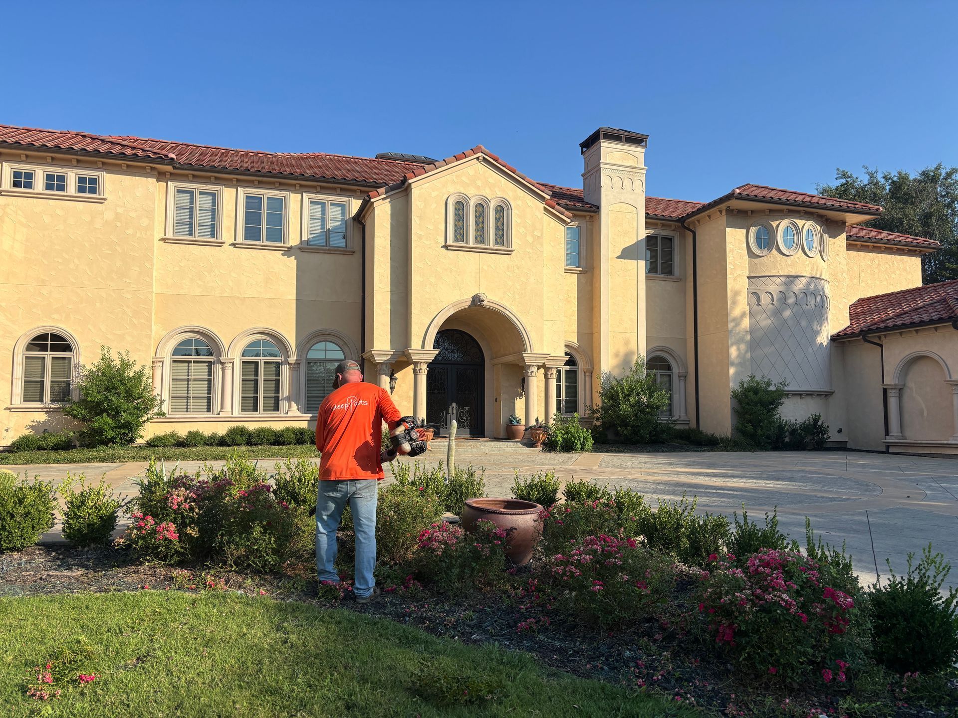 Person in orange jacket watering flowers in a sunny courtyard in front of a large cream-colored house