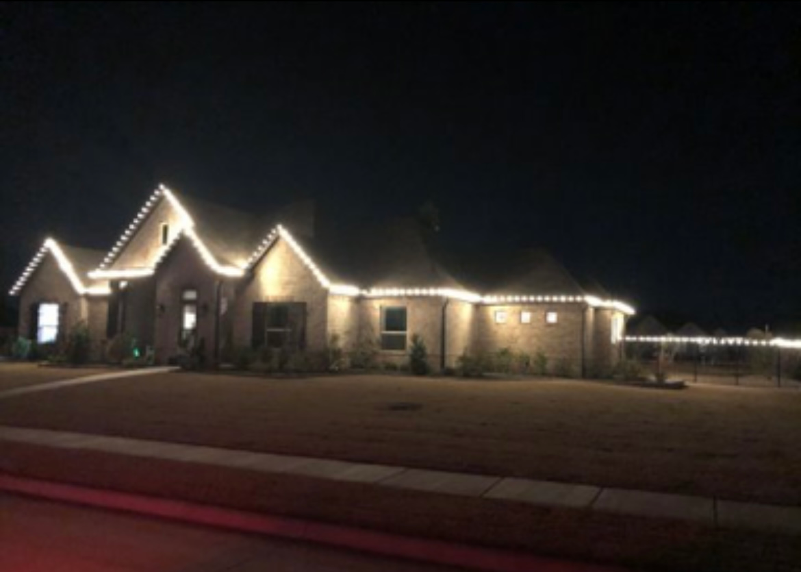 Single-story brick house at night, outlined with warm white holiday lights beside a dark street.