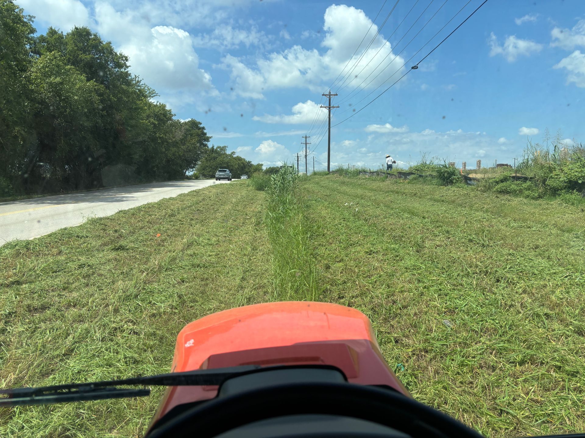 Rural road beside green field under blue sky, viewed from a red vehicle hood on a sunny day