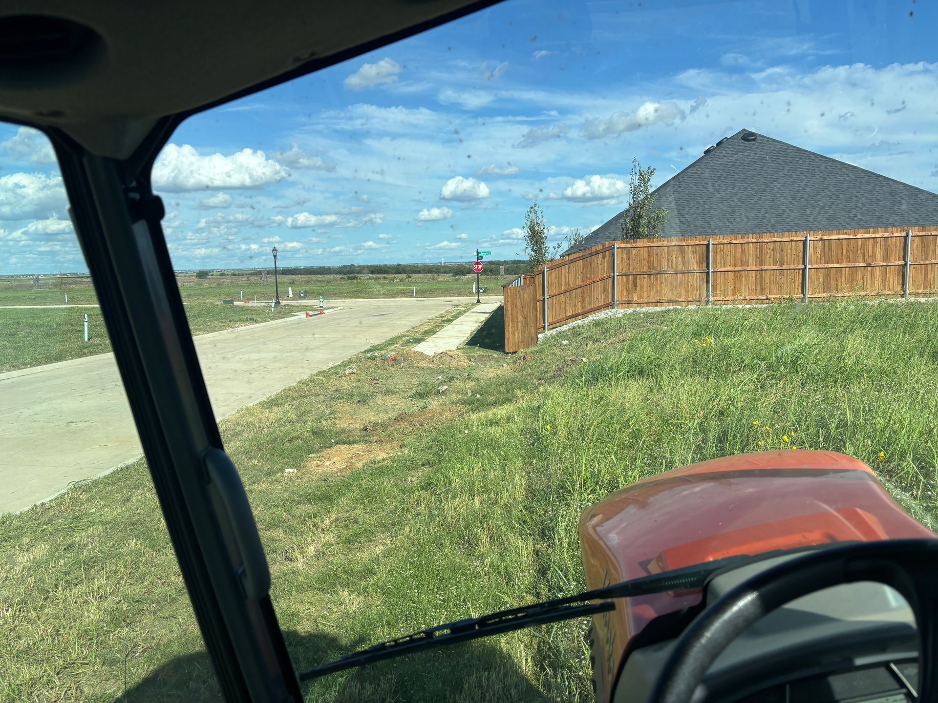 View from a vehicle over a grassy lot, sidewalk, and wooden fence beside a house on a sunny day