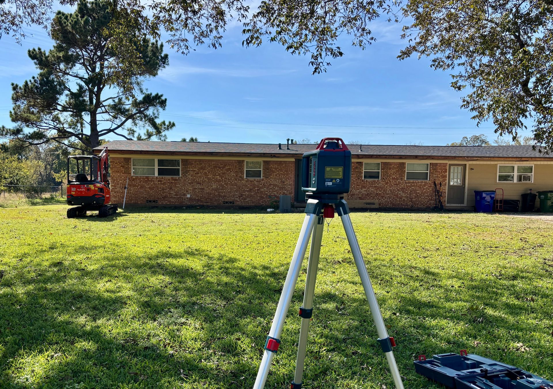 Surveying tripod on a sunny lawn in front of a brick building and trees