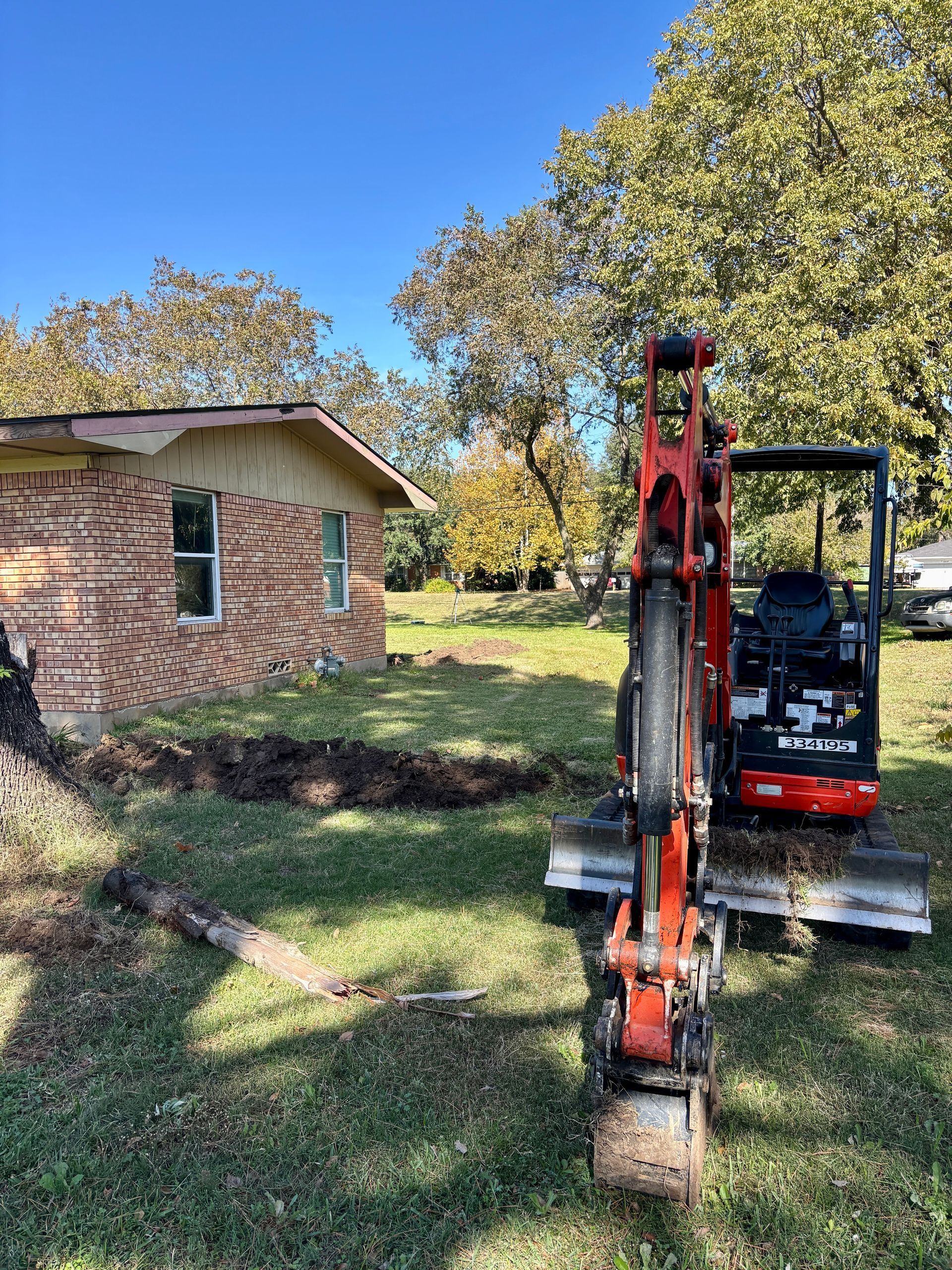 Small excavator parked beside a brick house in a grassy yard with autumn trees.