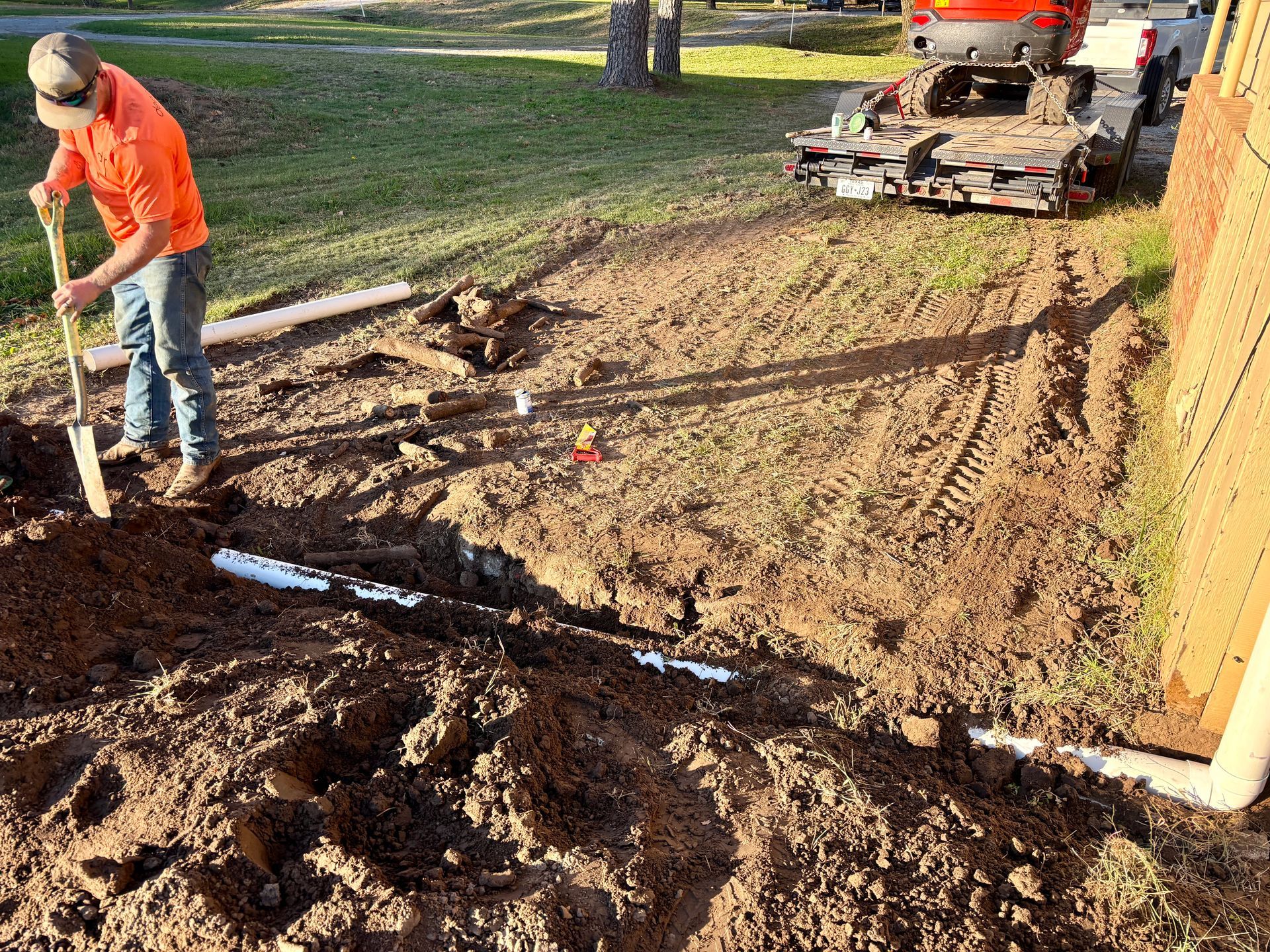 Worker shoveling mud beside a utility trench while a small excavator clears dirt nearby