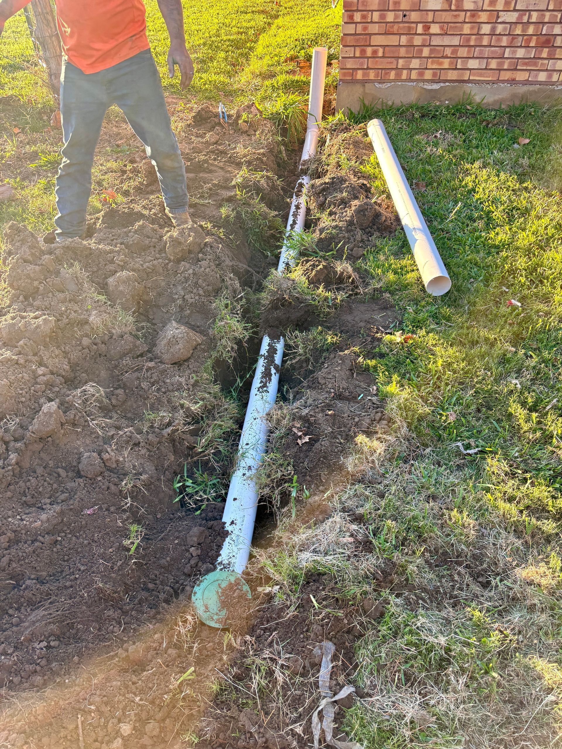 Worker beside dug trench with exposed white pipes in a grassy yard