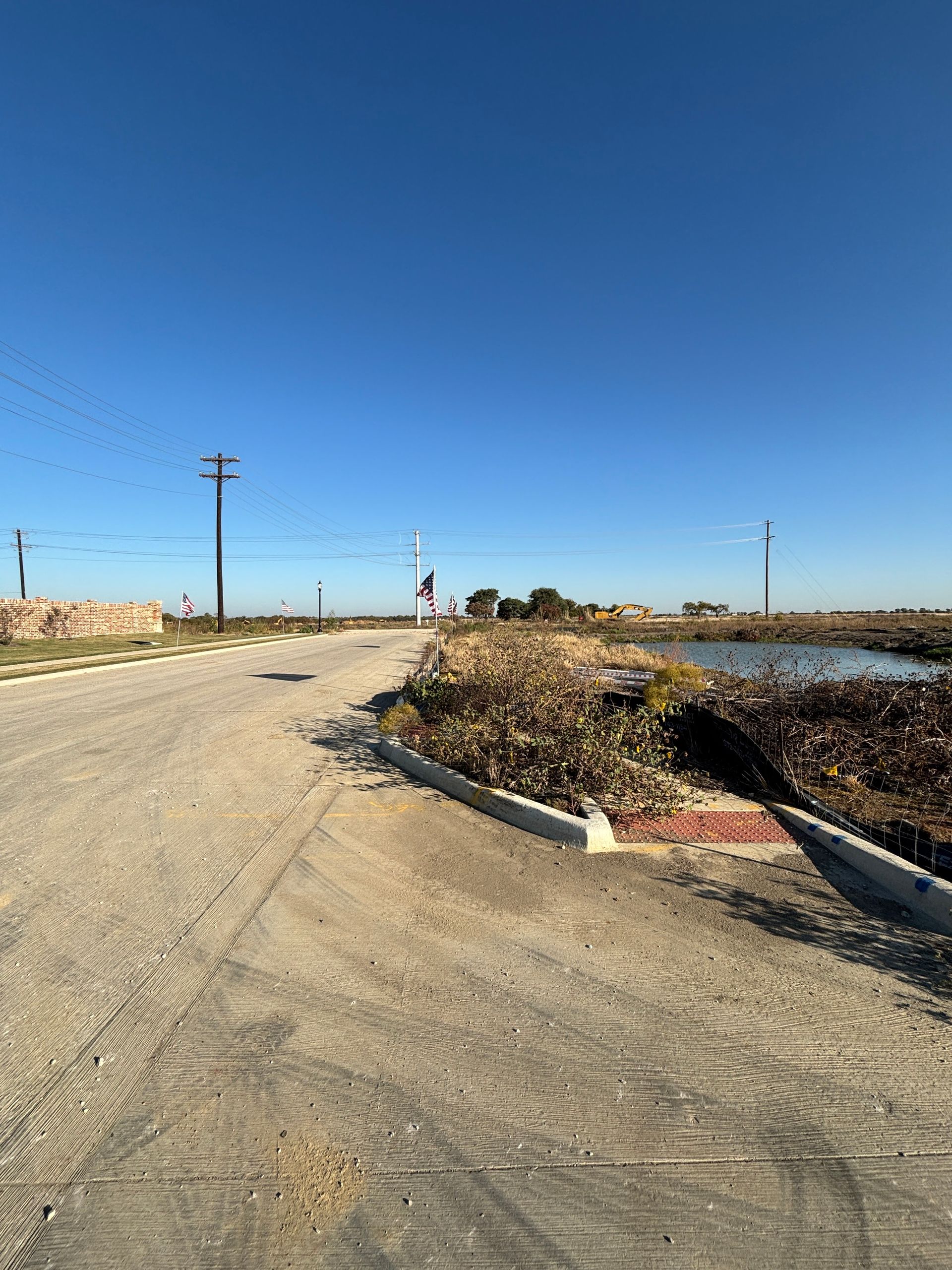 Rural dirt road beside a pond under a clear blue sky, with utility poles and dry brush.