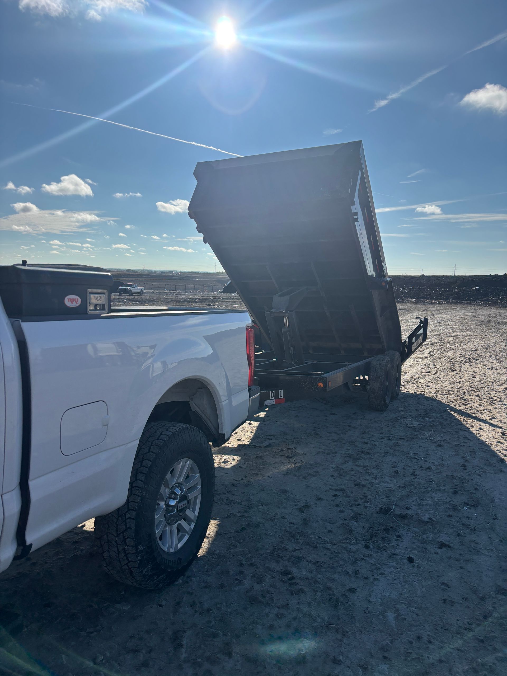 White pickup truck with its dump bed raised on a gravel lot under a bright blue sky