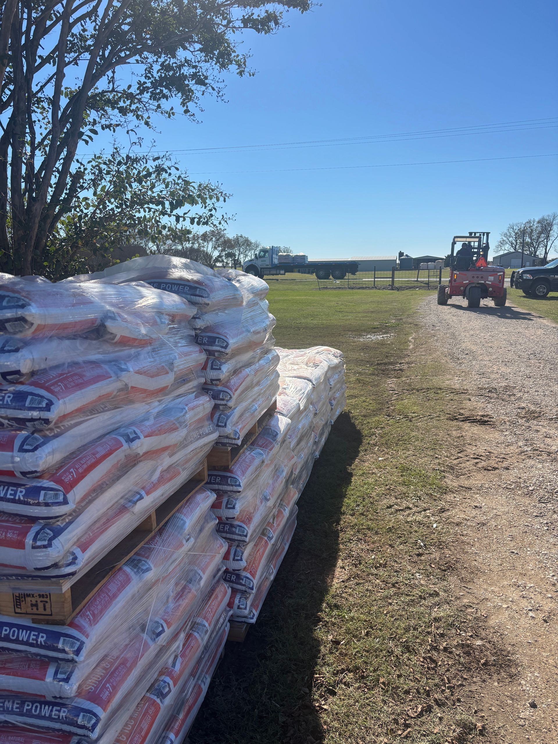 Stacks of bagged materials beside a gravel path, with a forklift in a sunny field in the distance.