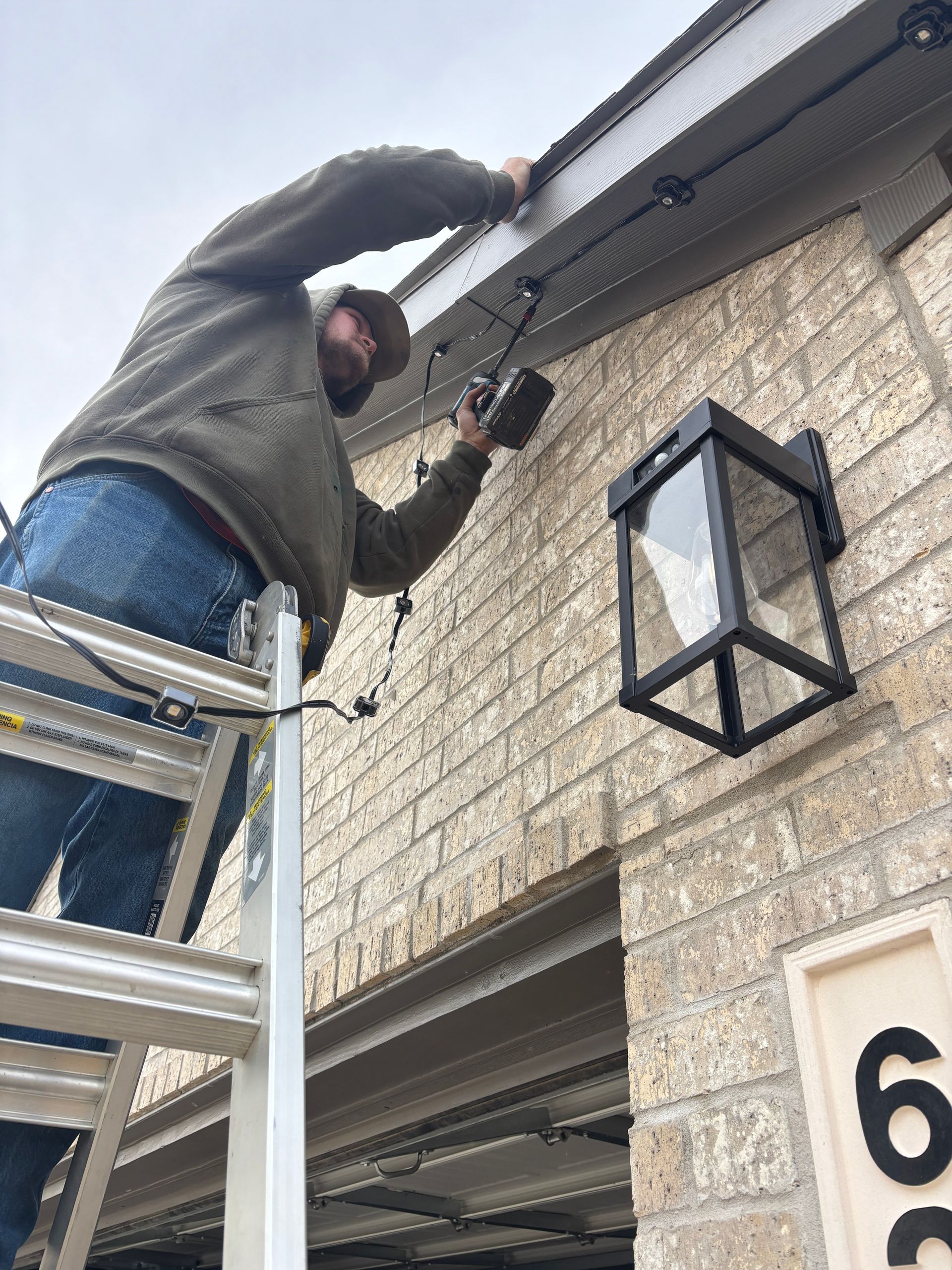 Worker on a ladder installing lights on a brick house exterior.