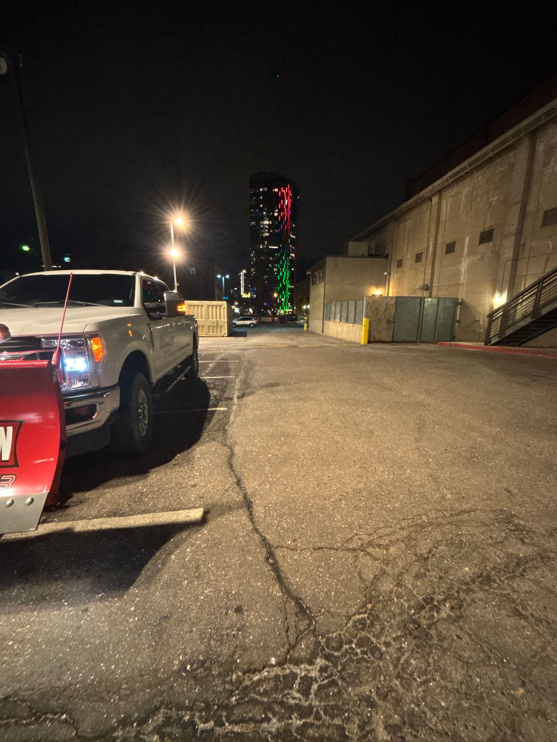 Nighttime industrial lot with parked trucks, a lit high-rise in the distance, and a cracked paved lane.