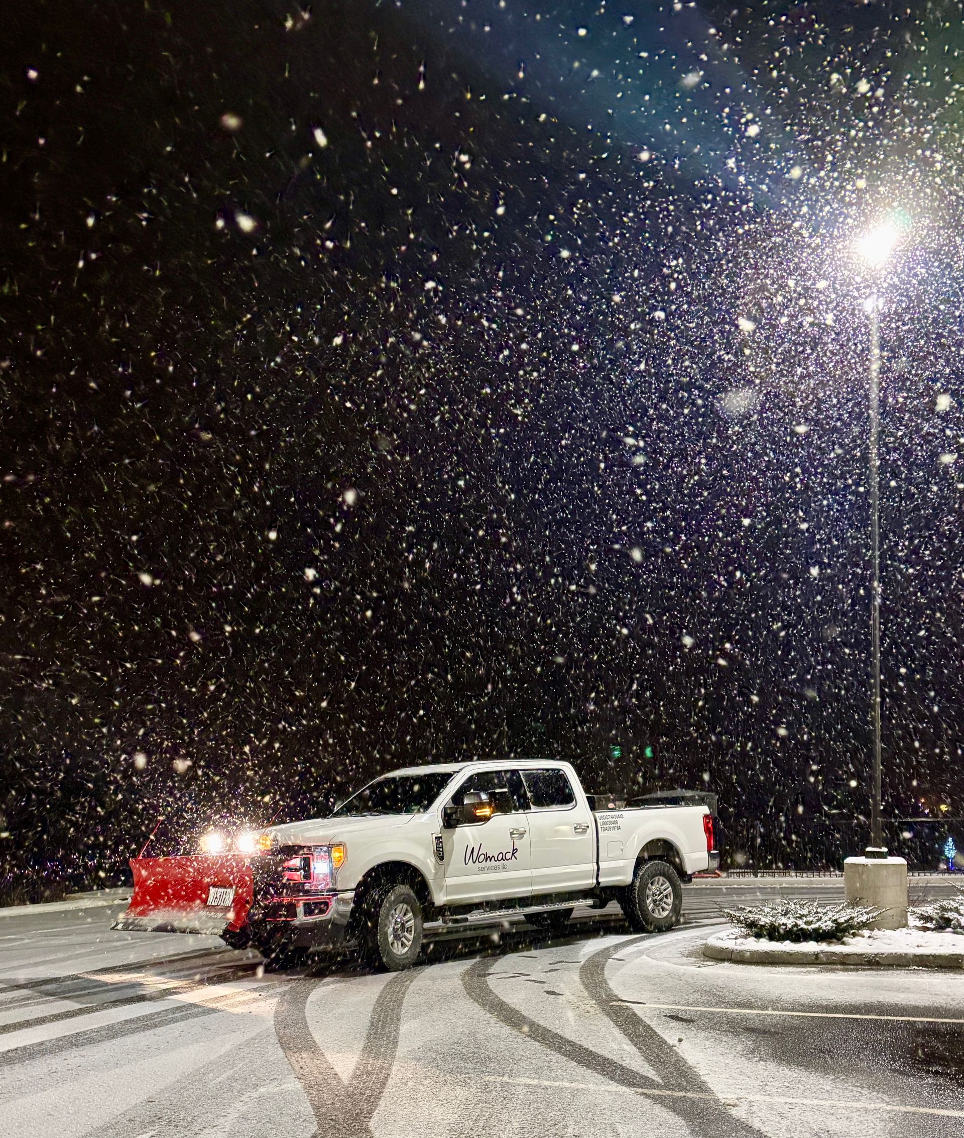 White pickup truck in snowy night parking lot, headlights on, with falling snow and tire tracks.