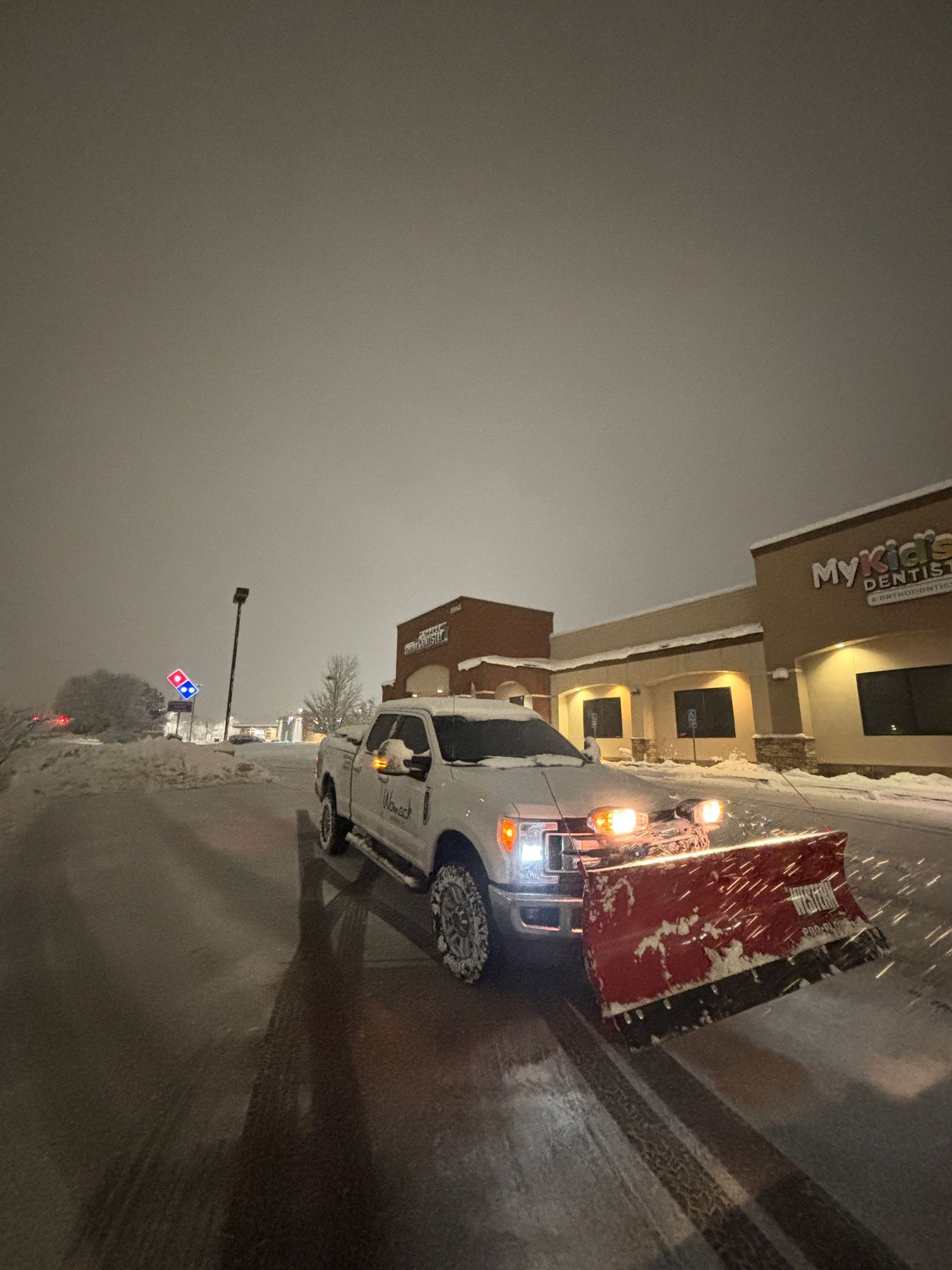Snowplow clearing a snowy street beside a building at night, with a raised red plow blade.