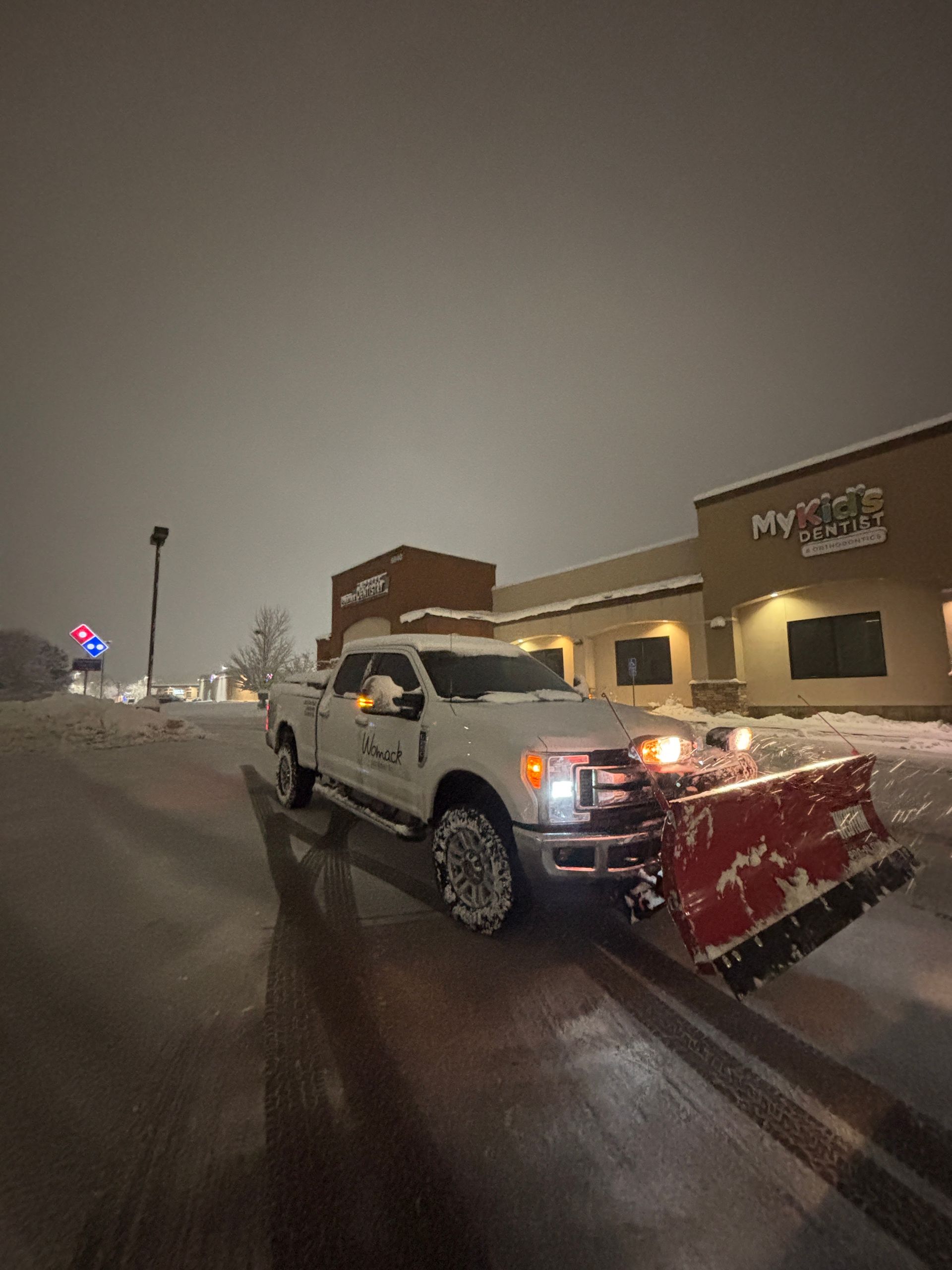 White pickup truck with red snowplow on a snowy street beside a lit building at night