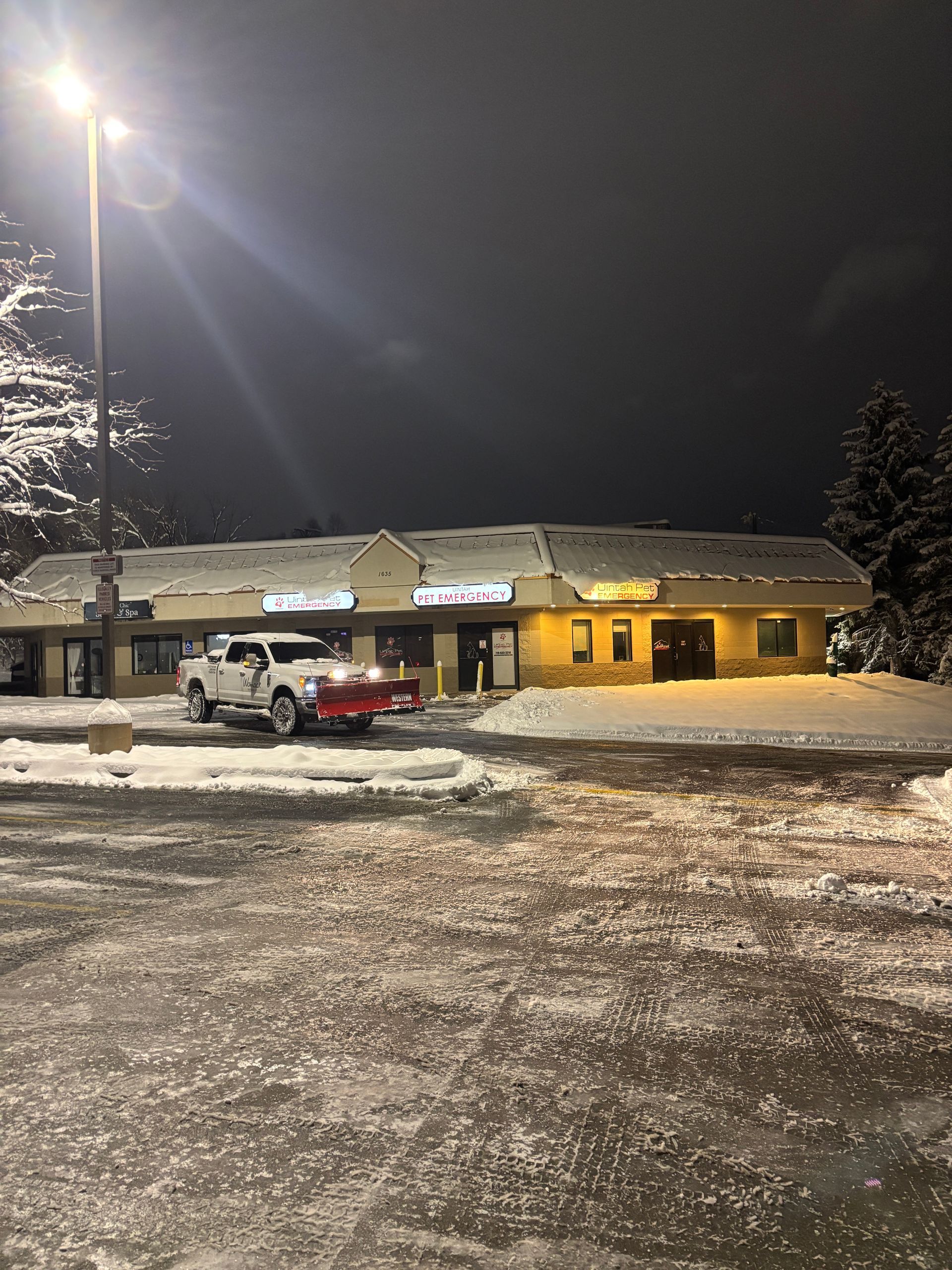 Snowy parking lot outside a lit building at night, with a pickup truck and streetlights.