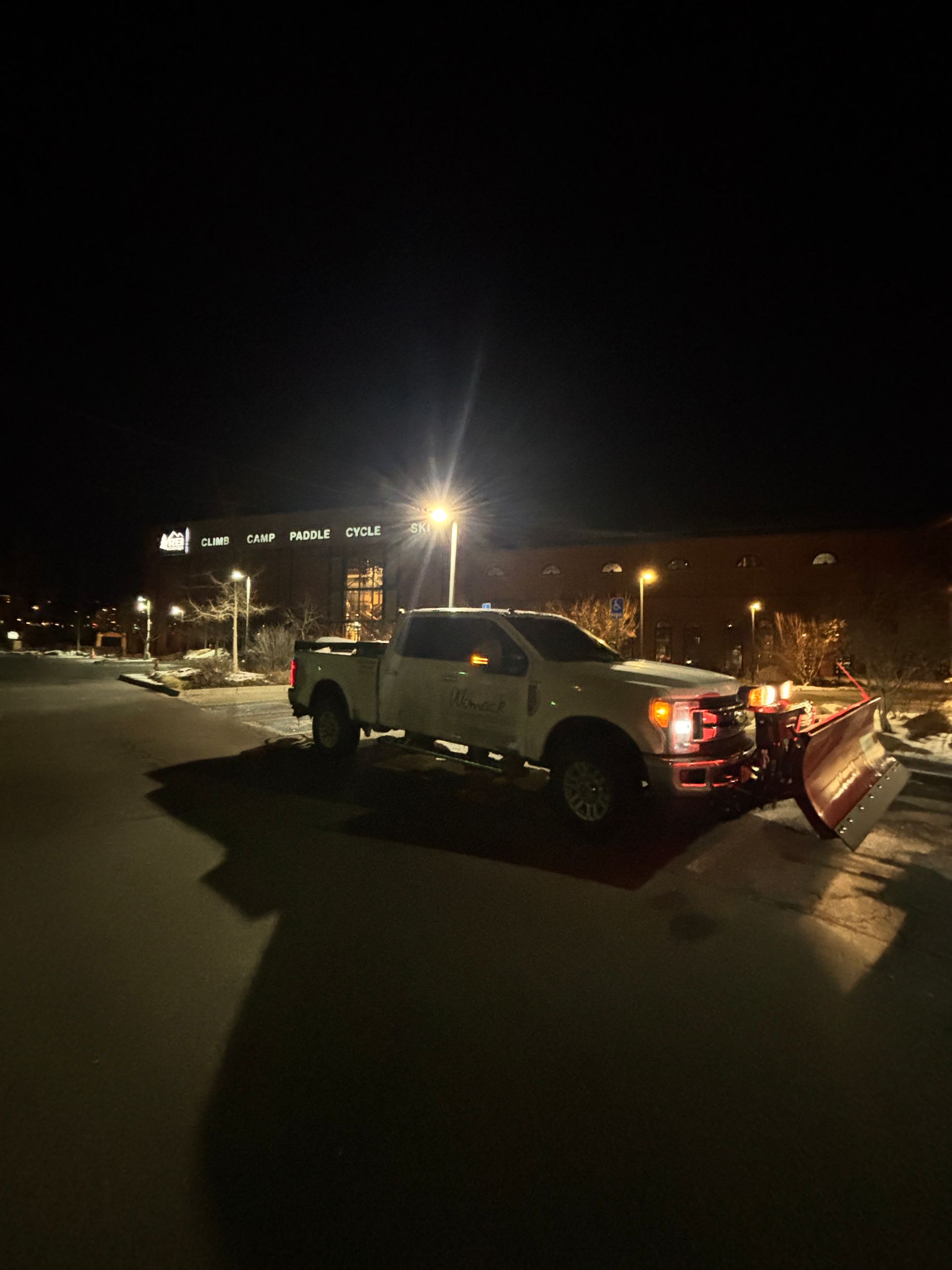 Tow truck with plow attachment parked on a snowy street at night under bright lights
