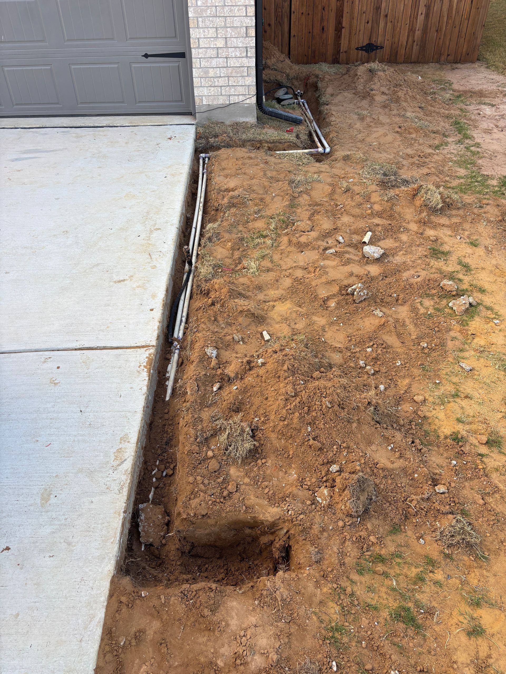 Concrete sidewalk beside dirt yard with an open trench along the edge near a garage door.