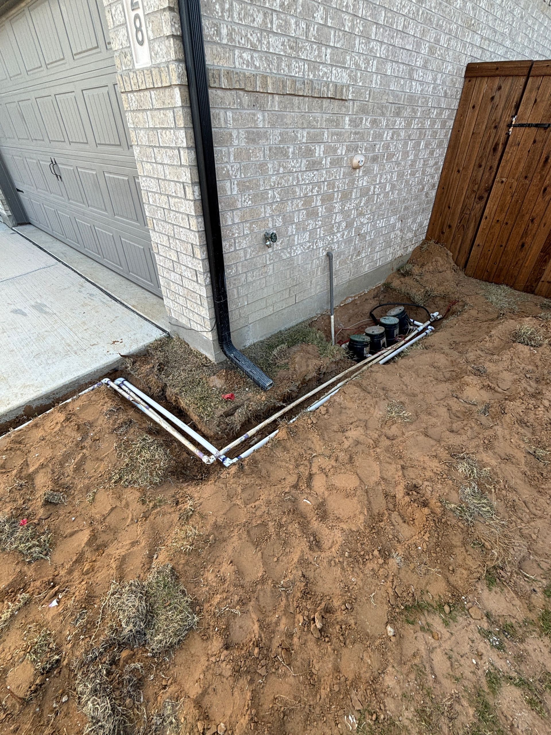 Exterior corner of a house with exposed drainage pipes and dirt around the foundation