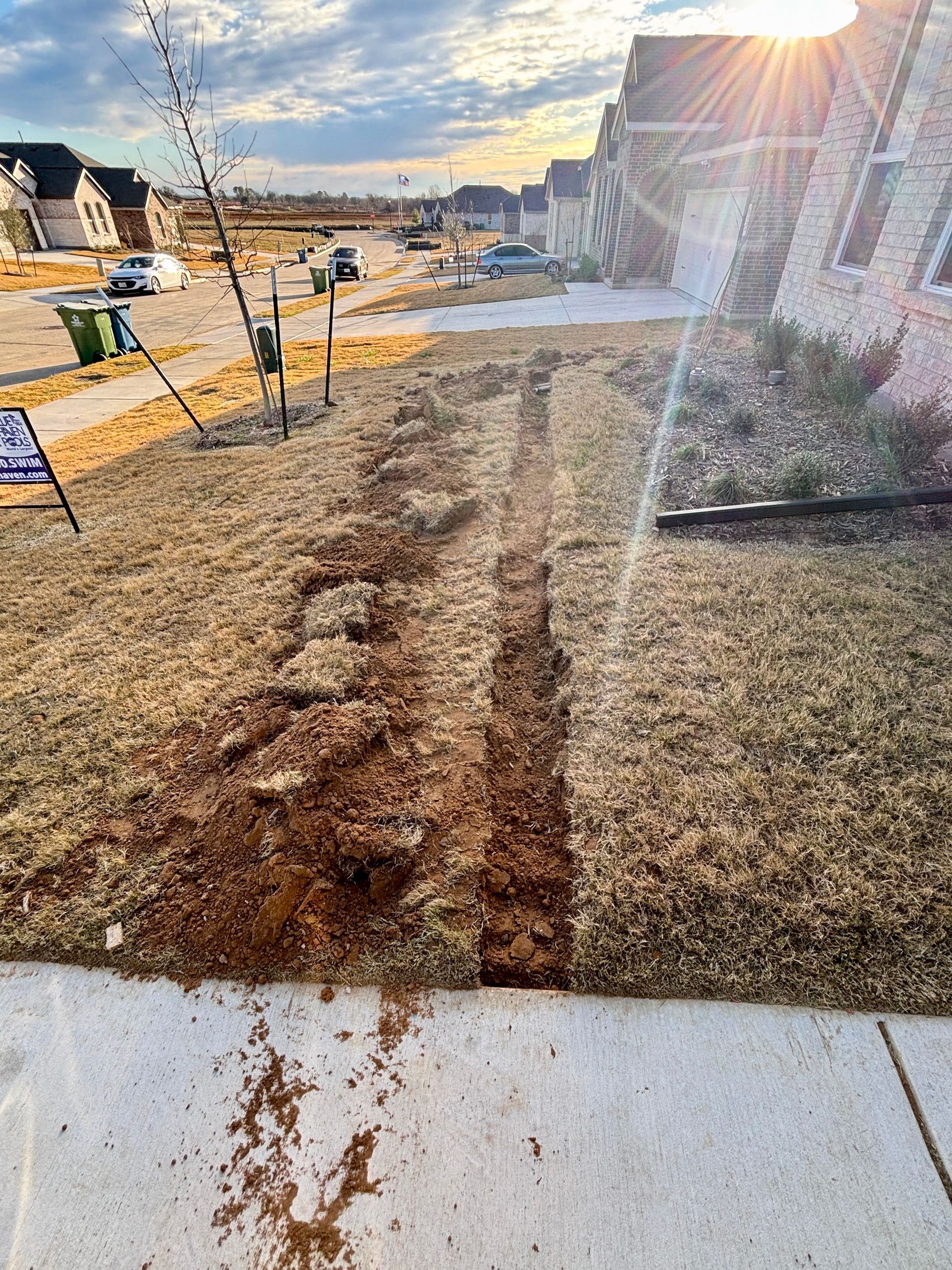 Sunlit residential street with a muddy dirt path beside a sidewalk and houses under construction